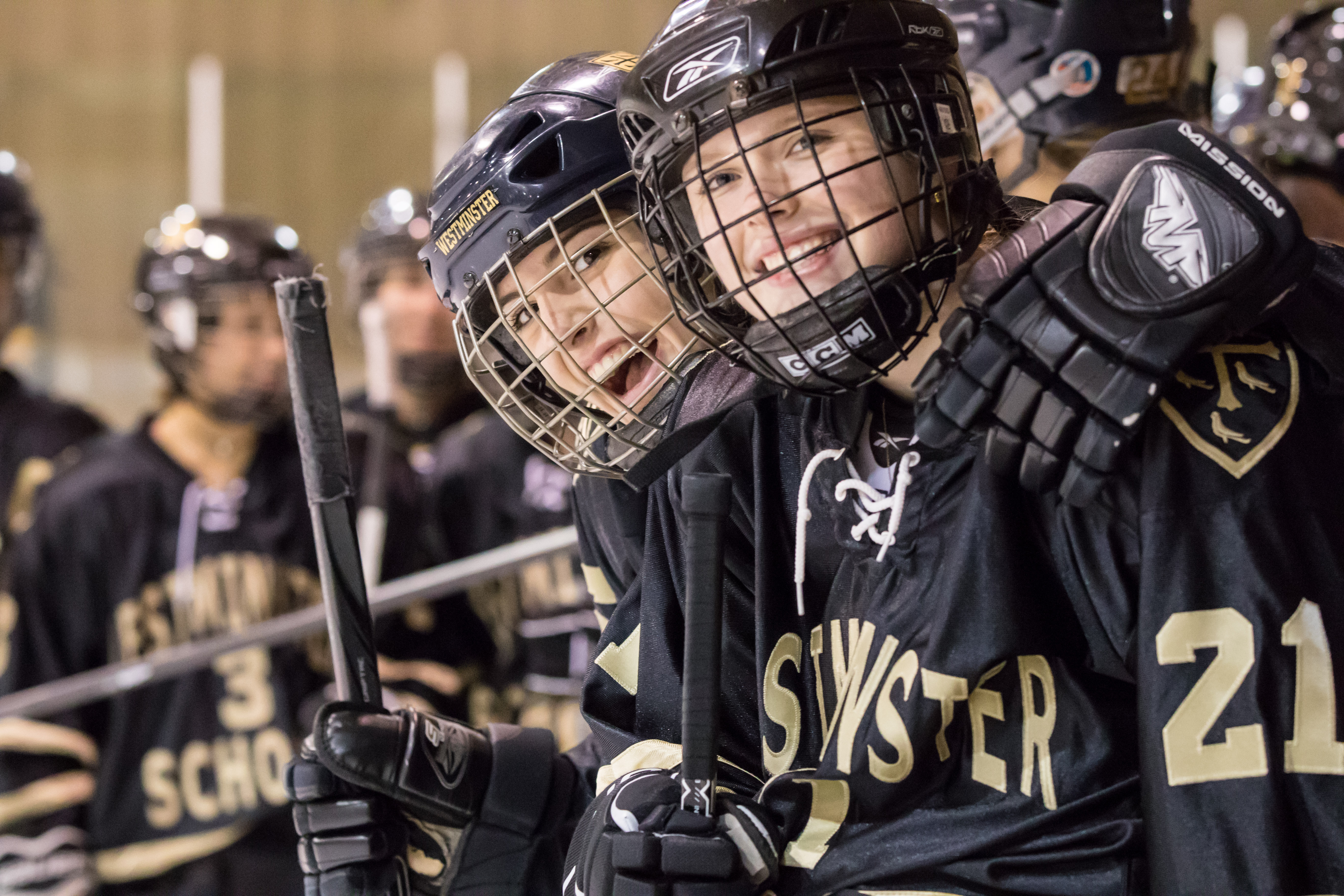 Two smiling youth hockey players in black jerseys and helmets stand at a rink bench.