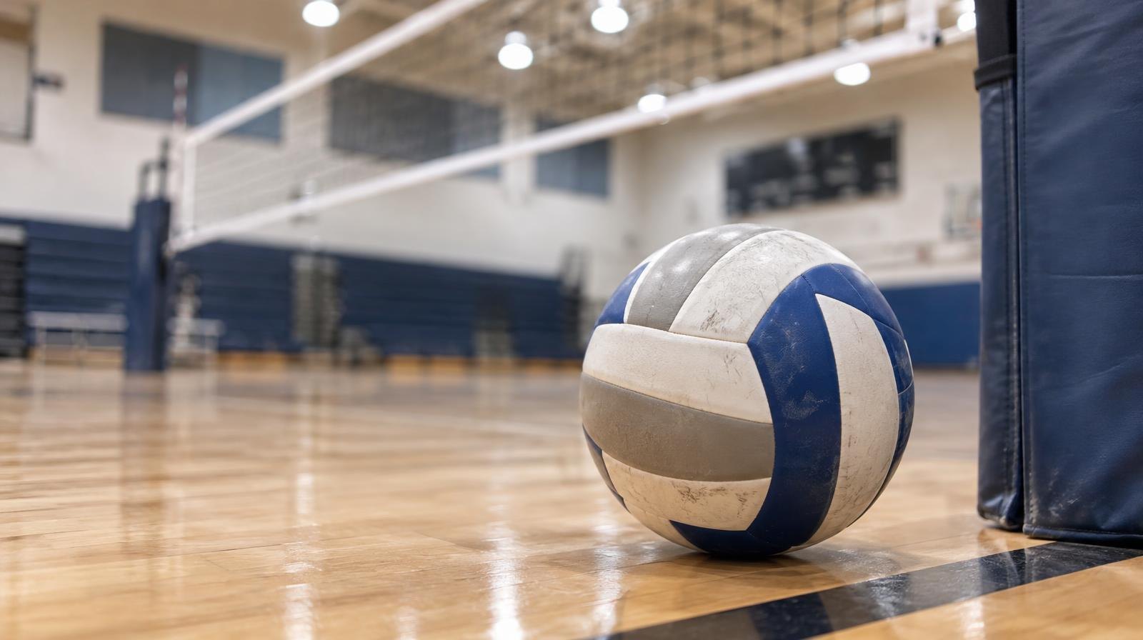 A volleyball resting on a hardwood gym floor near a net post.