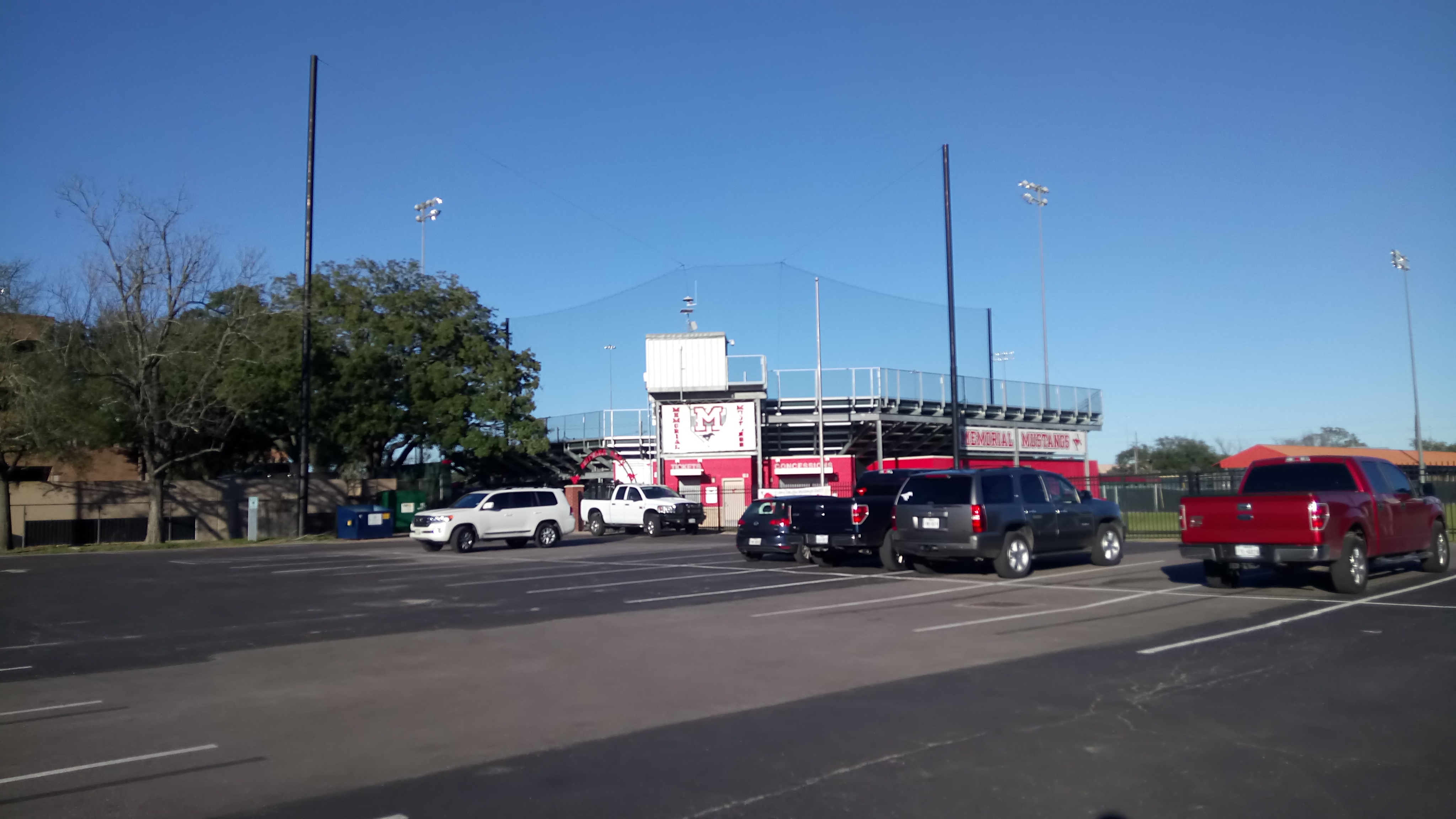 High school baseball stadium exterior with light poles, bleachers, and a scoreboard under blue sky.