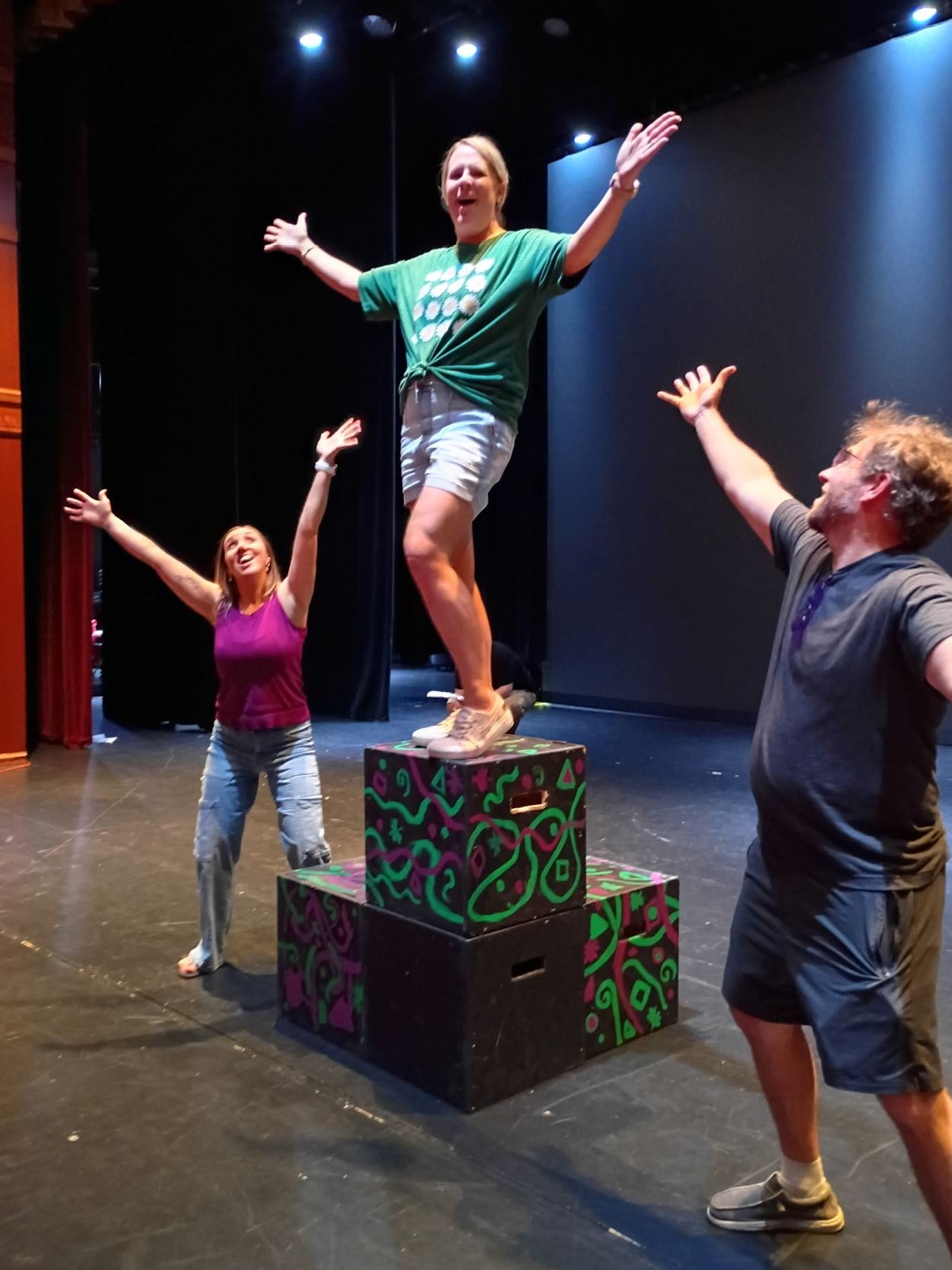 Three performers on a stage with arms outstretched near colorfully painted boxes.