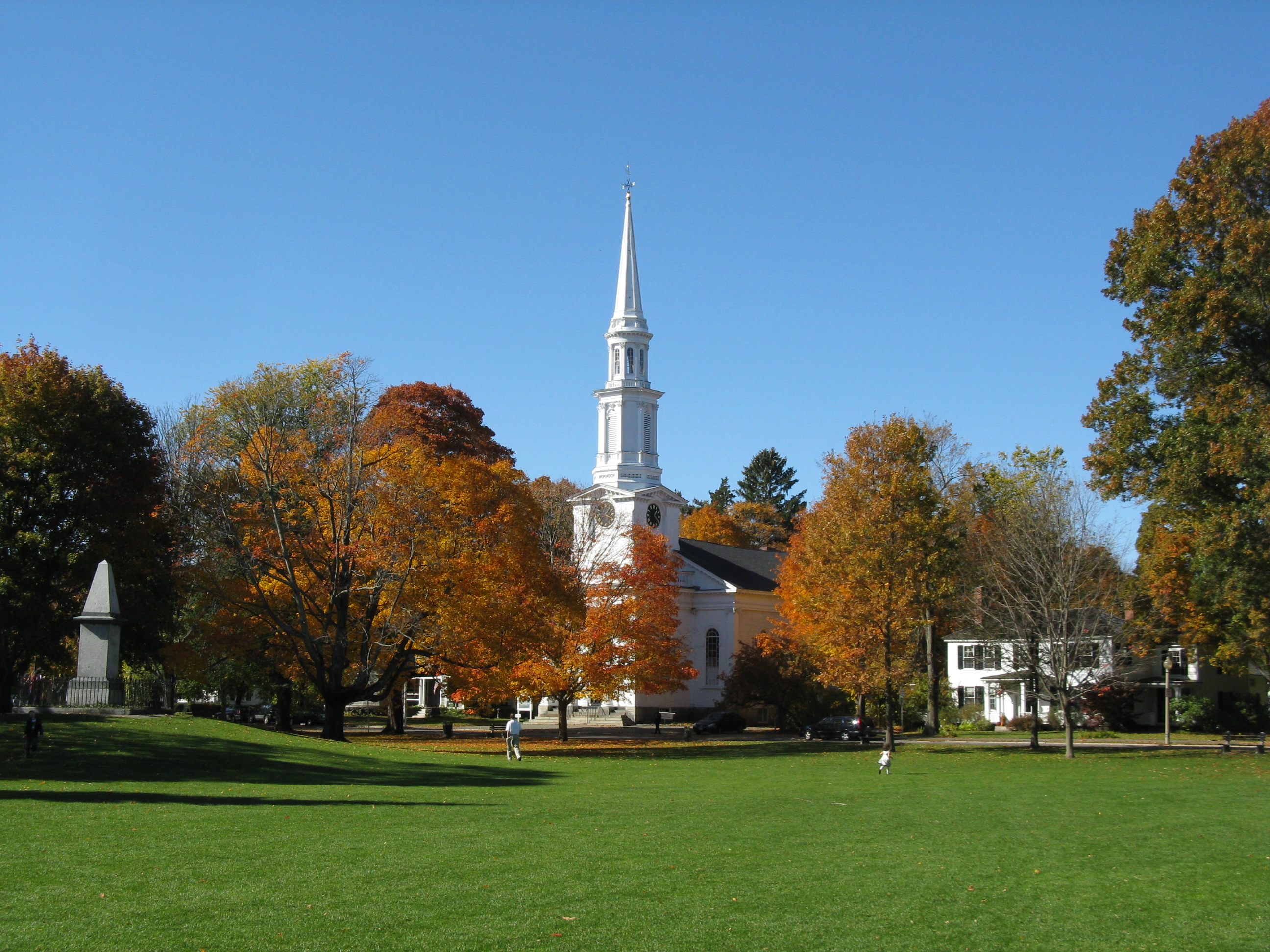 White colonial church and obelisk monument on a tree-lined town green in autumn.