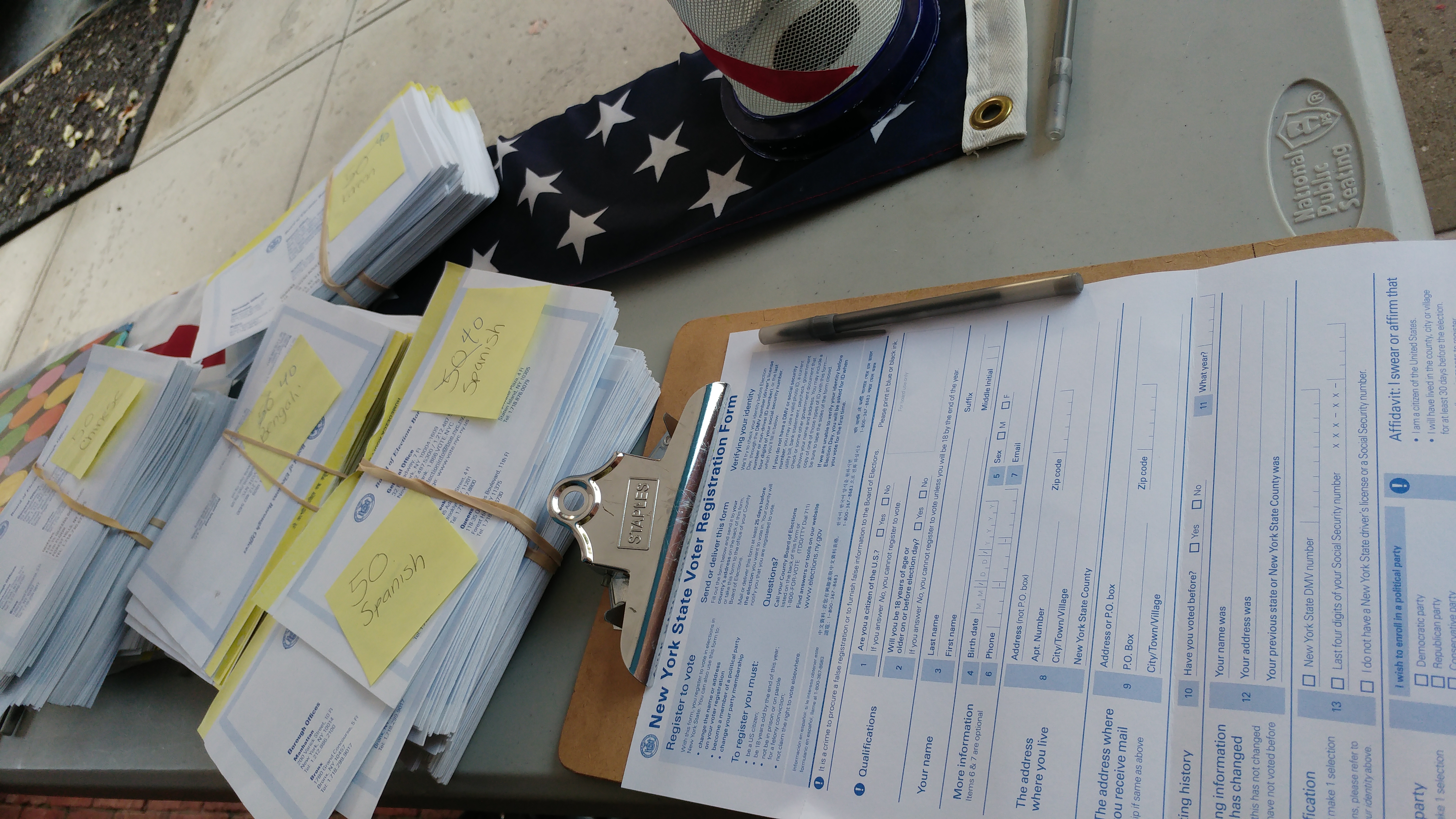 Voter registration form on a clipboard with a pen, beside stacks of multilingual forms and a US flag.