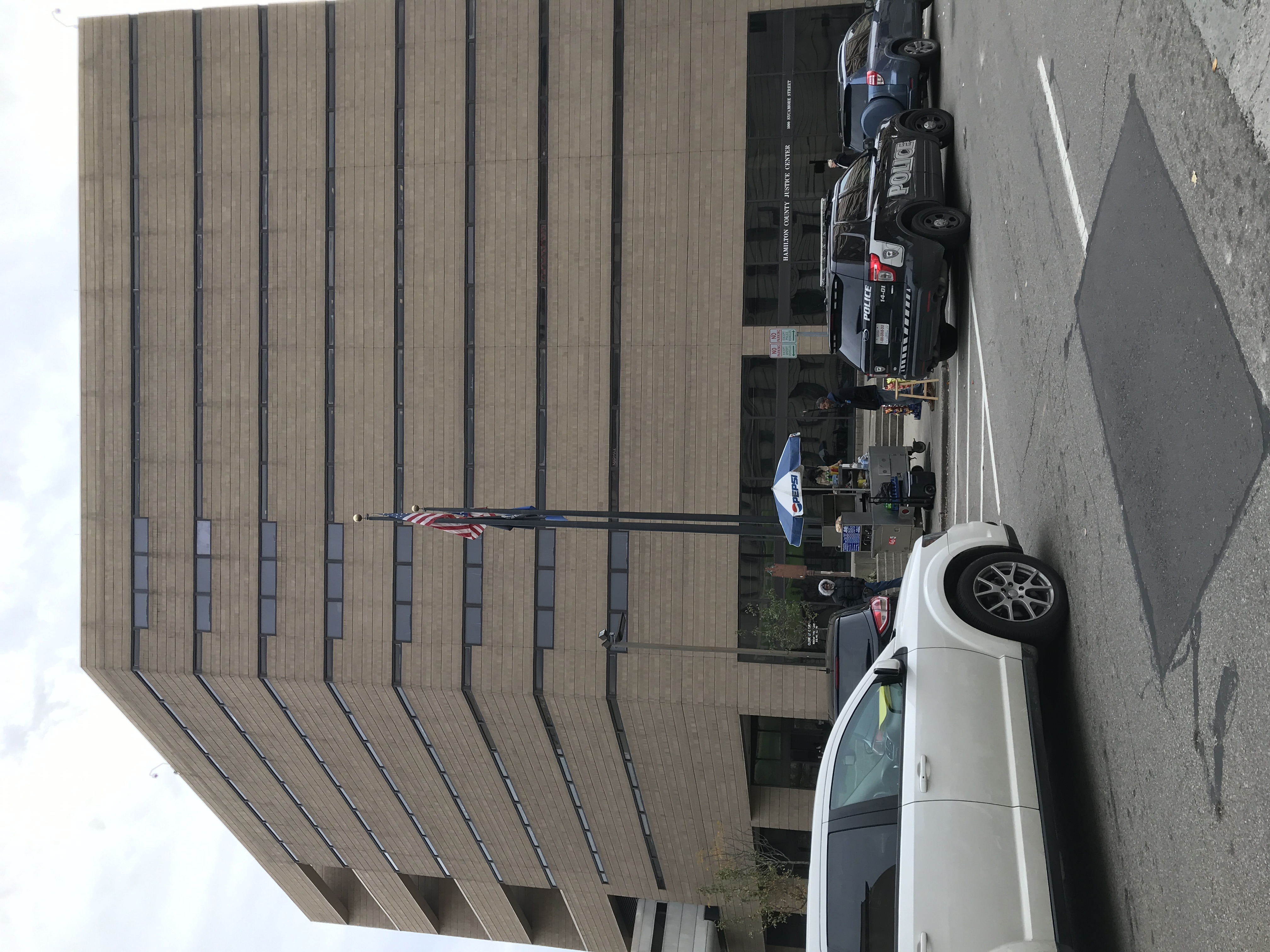 Multi-story government building with police vehicles parked out front and an American flag on a flagpole.