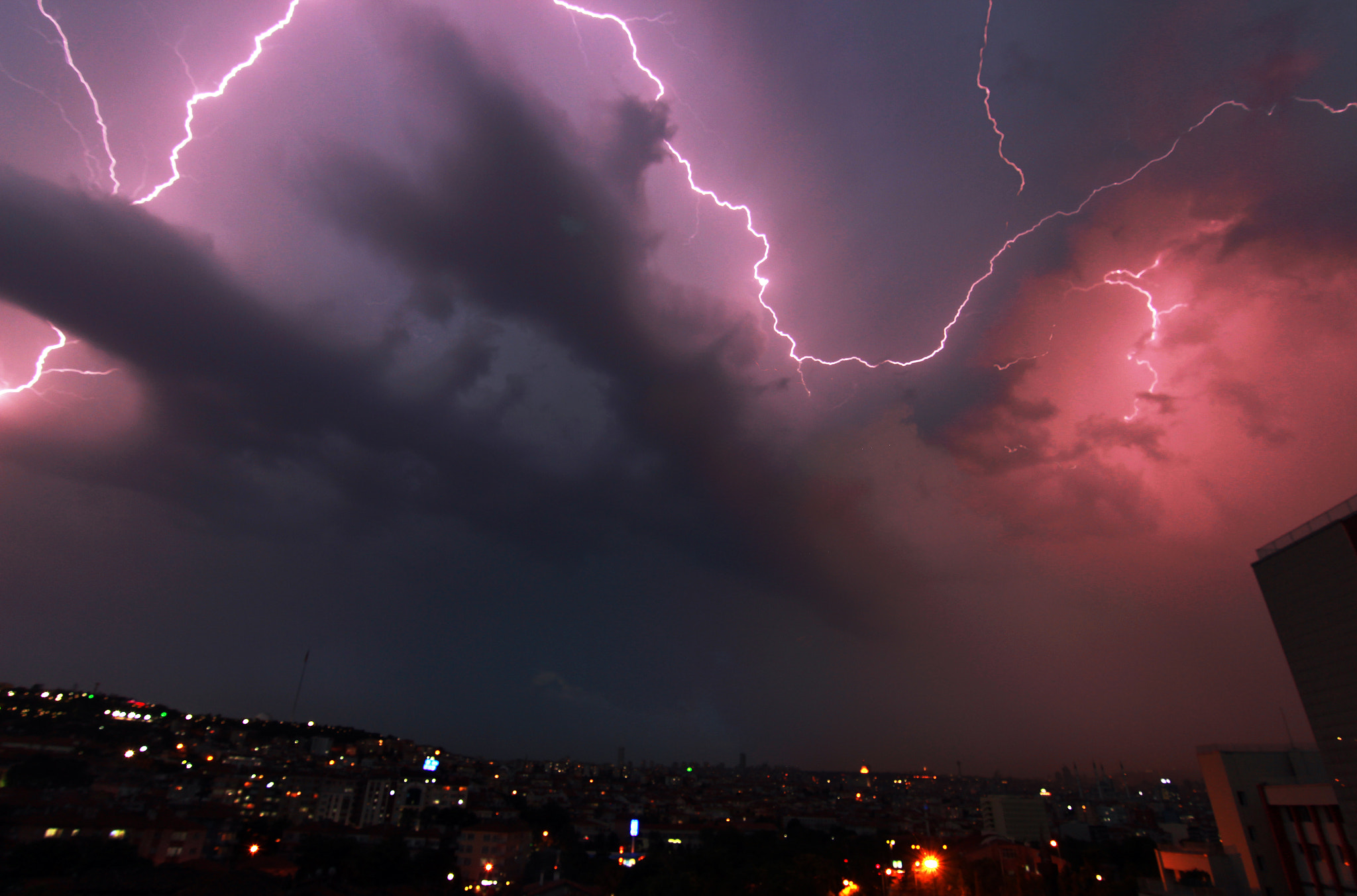 Multiple lightning bolts illuminate dark storm clouds over a city at night.