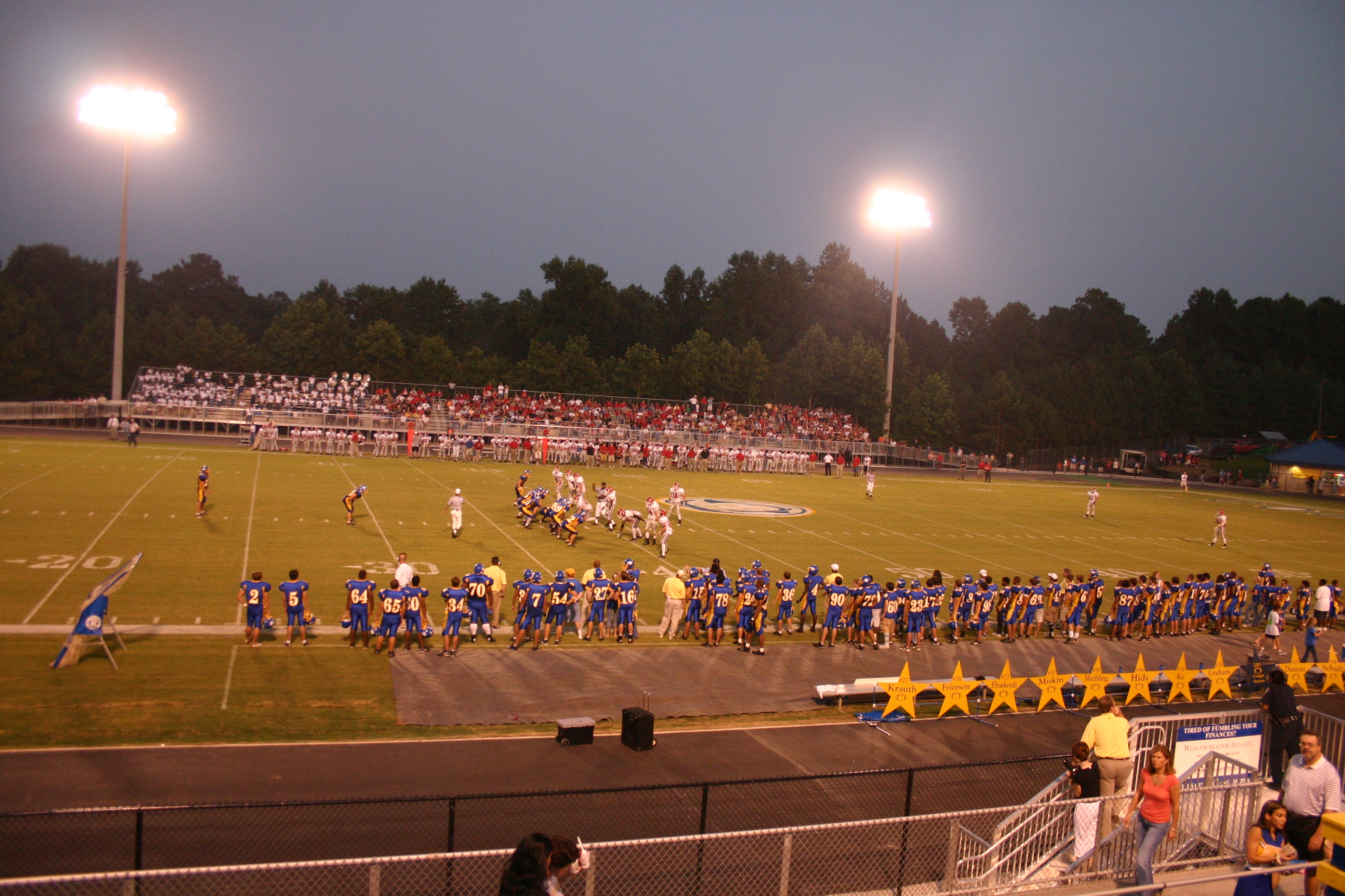 High school football players on a lit field at dusk with packed bleachers.