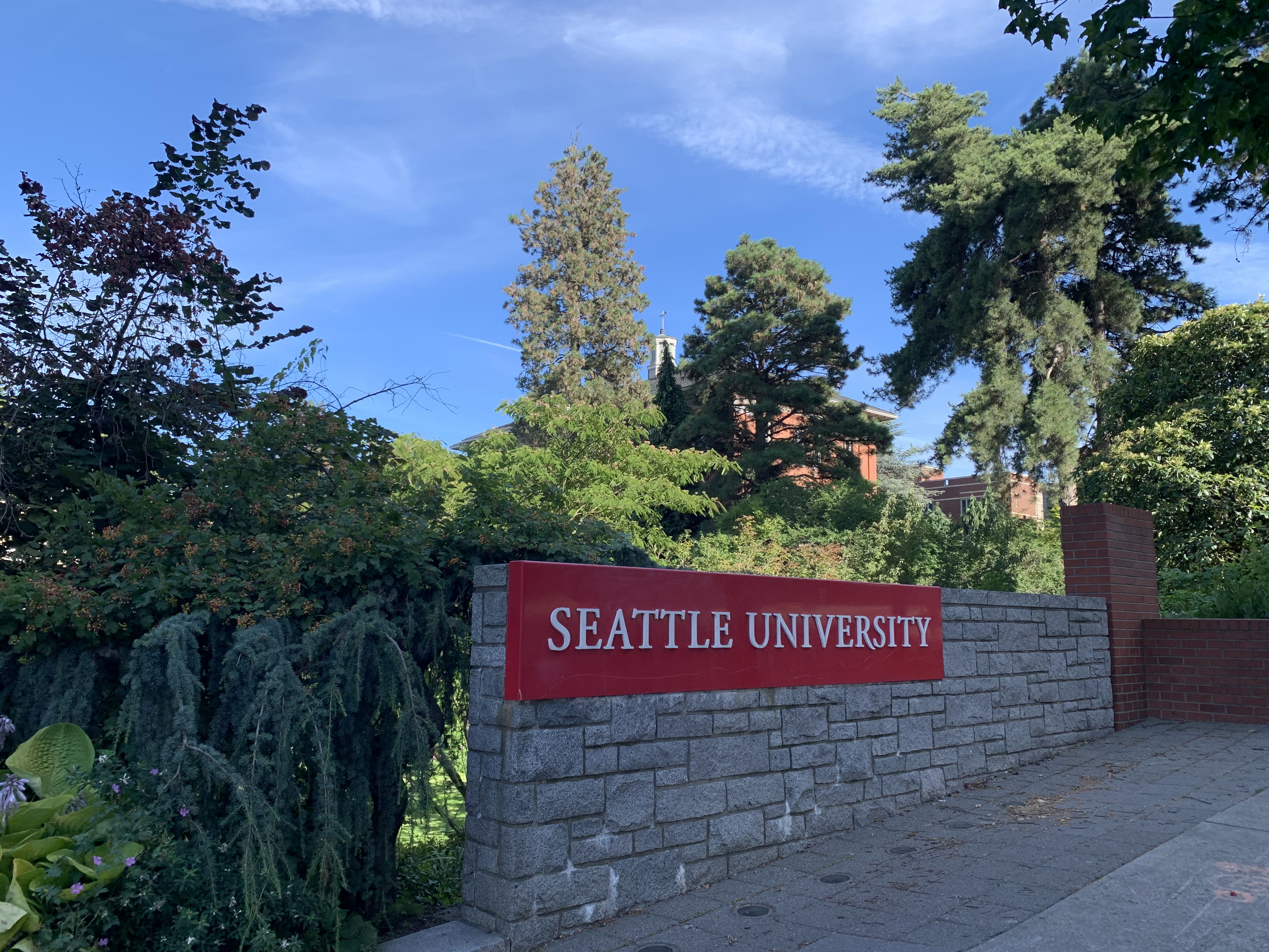 Red Seattle University sign on a stone wall near a tree-lined campus entrance.
