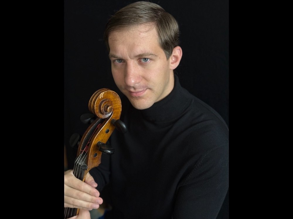 A man in a black turtleneck holding the scroll and neck of a wooden cello against a dark background.