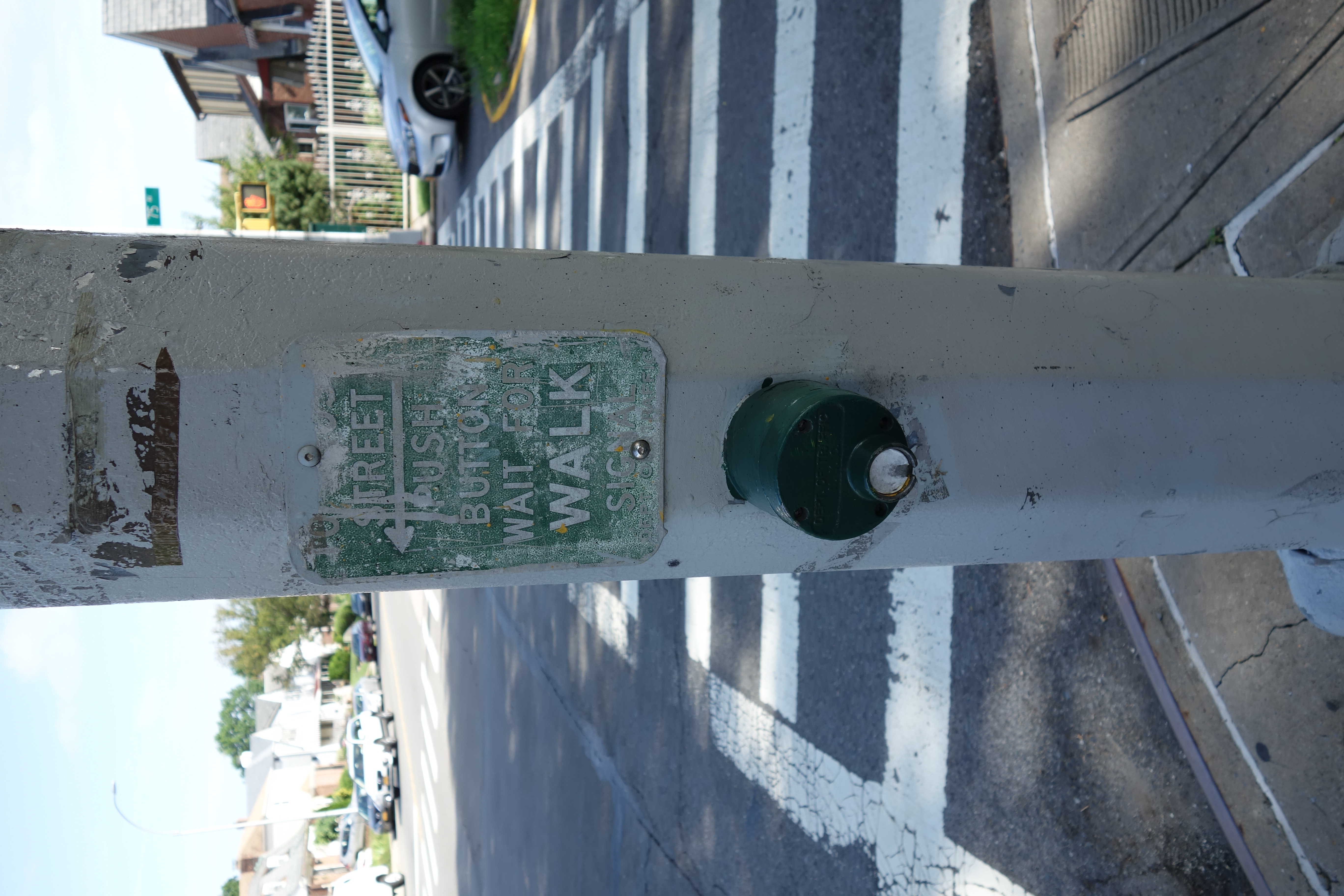 Weathered green dome-shaped pedestrian push button on a white pole at a street corner crosswalk.