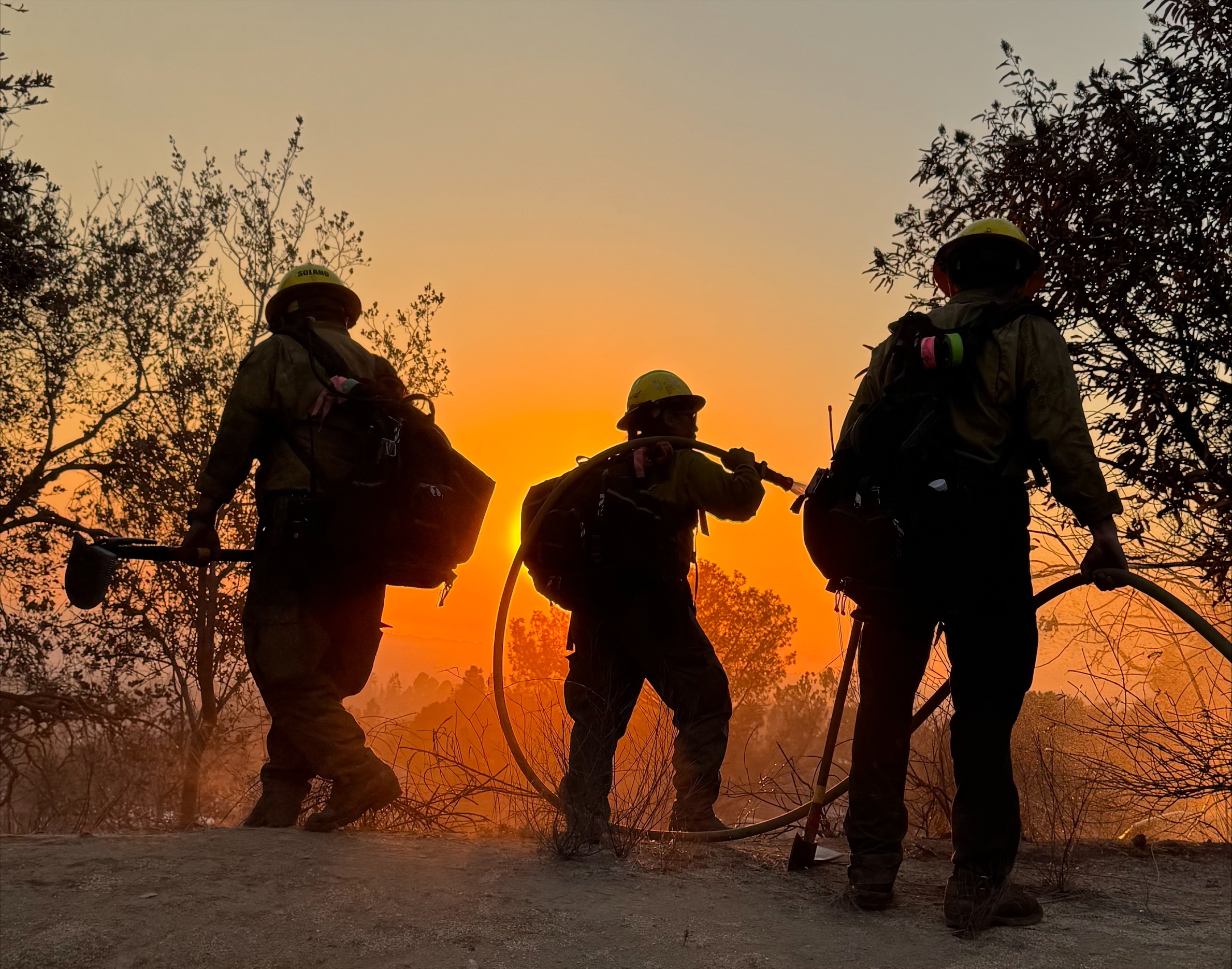 Three firefighters in silhouette against an orange smoke-filled sky carrying fire hoses.