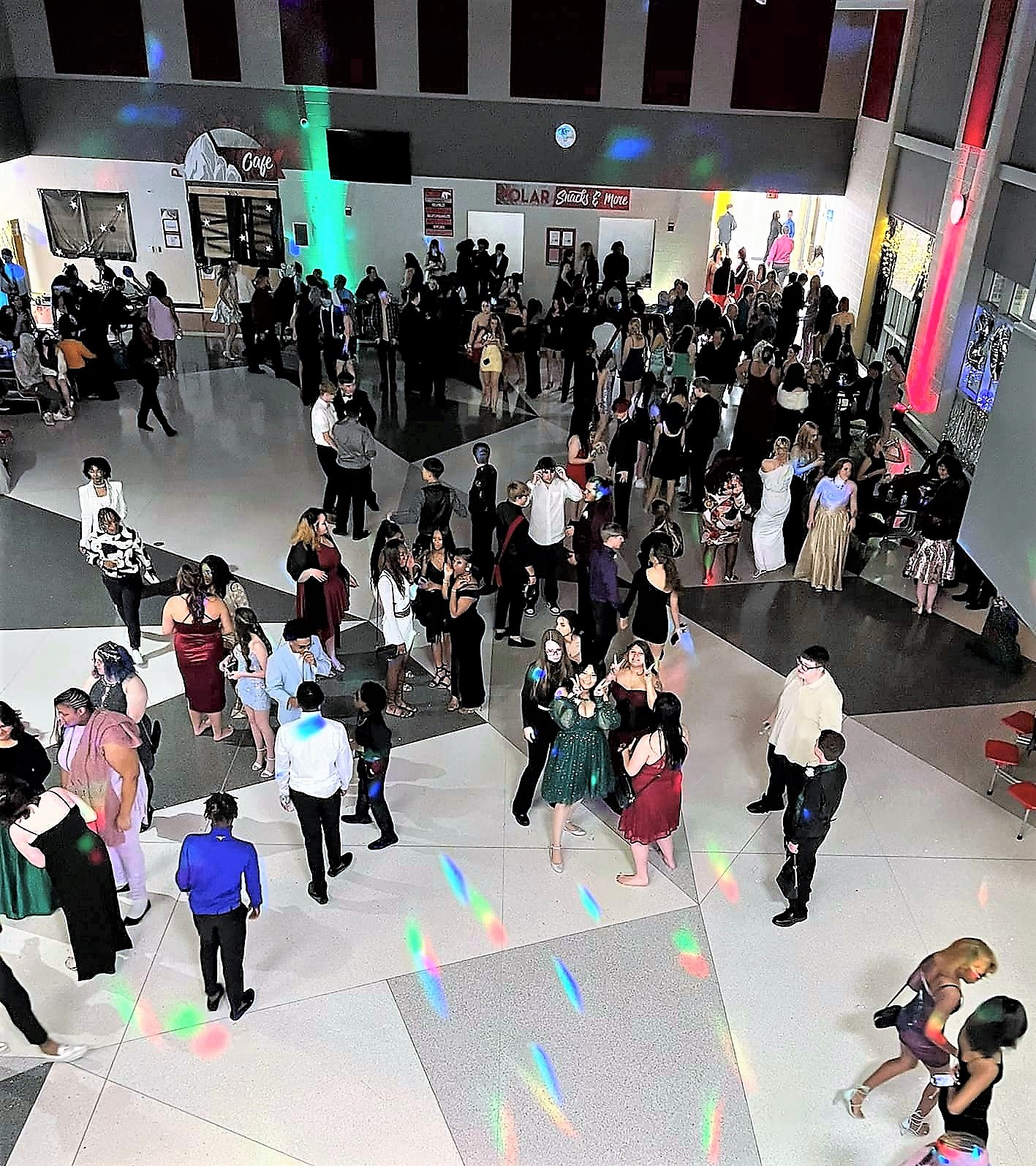 Students in formal attire dancing on a colorfully lit dance floor inside a school building.