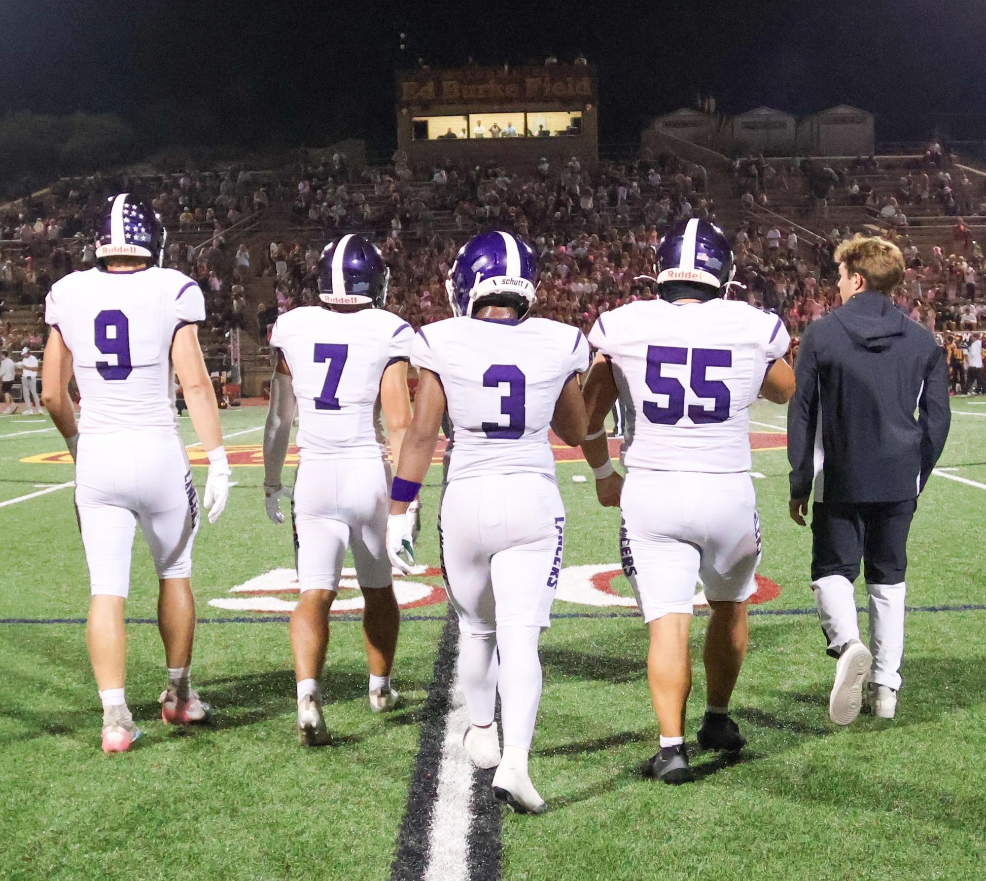 Four football players in white and purple uniforms walk toward a packed stadium at night.