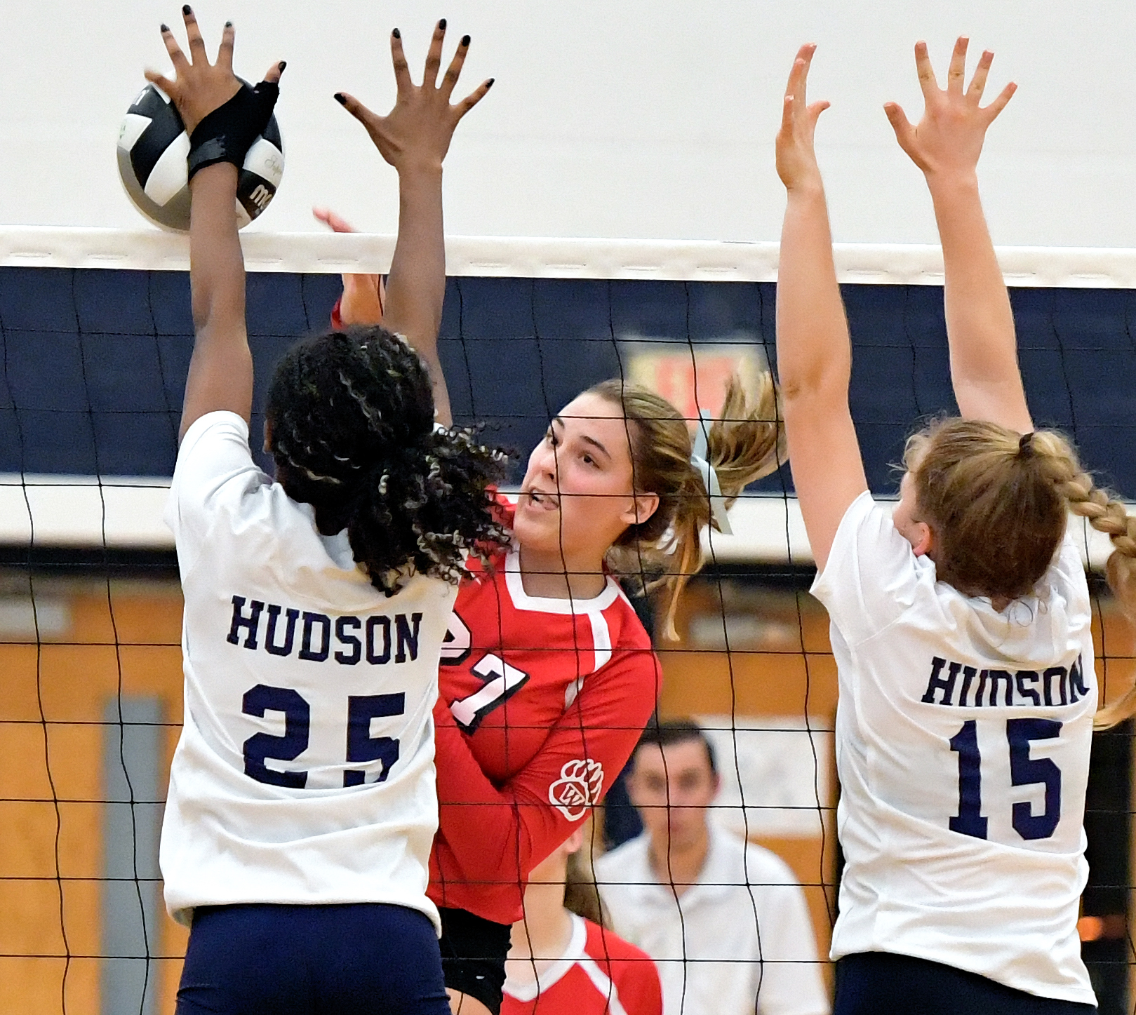 A volleyball player in red spikes the ball as two players in white attempt a block at the net.