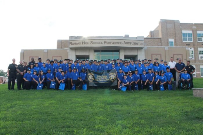 Large group of young people in blue uniforms and caps posed outdoors in front of a performing arts center.