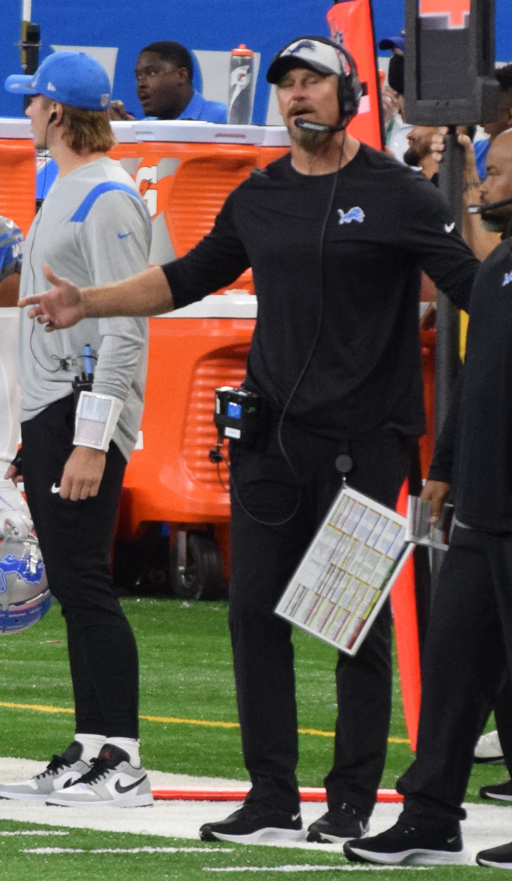 Man in Detroit Lions gear wearing a headset and holding a play sheet on the sideline.