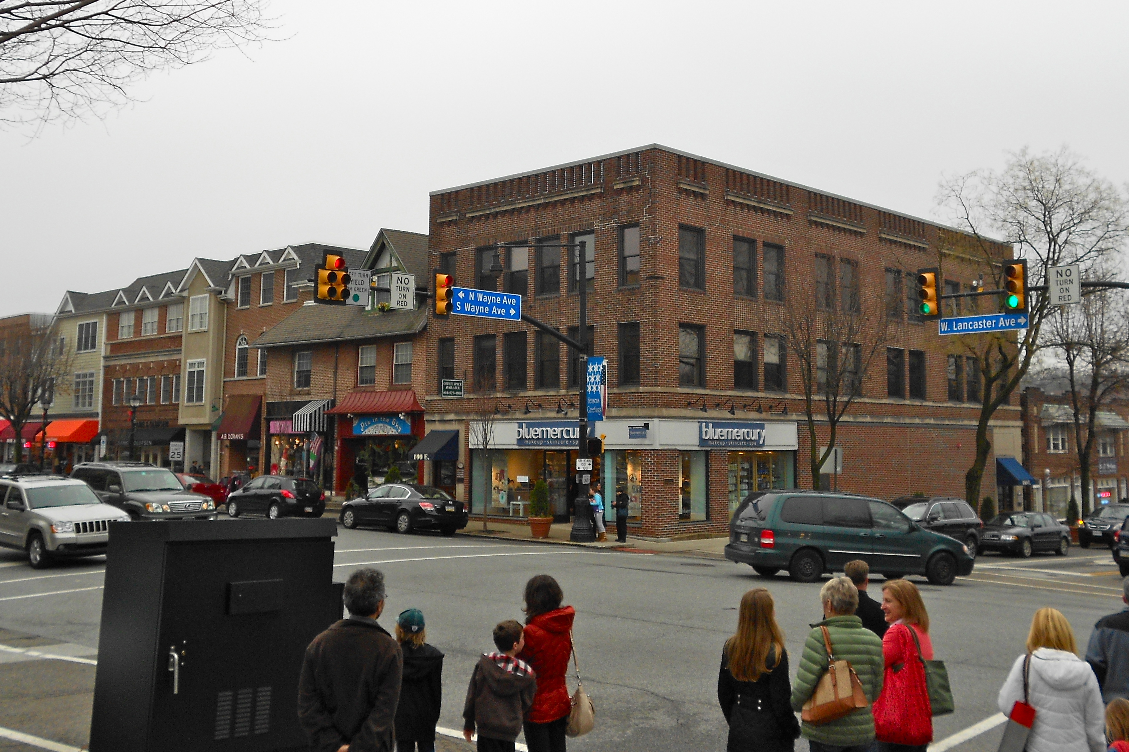 Street-level view of a busy commercial intersection with brick storefronts and pedestrians at a crosswalk.