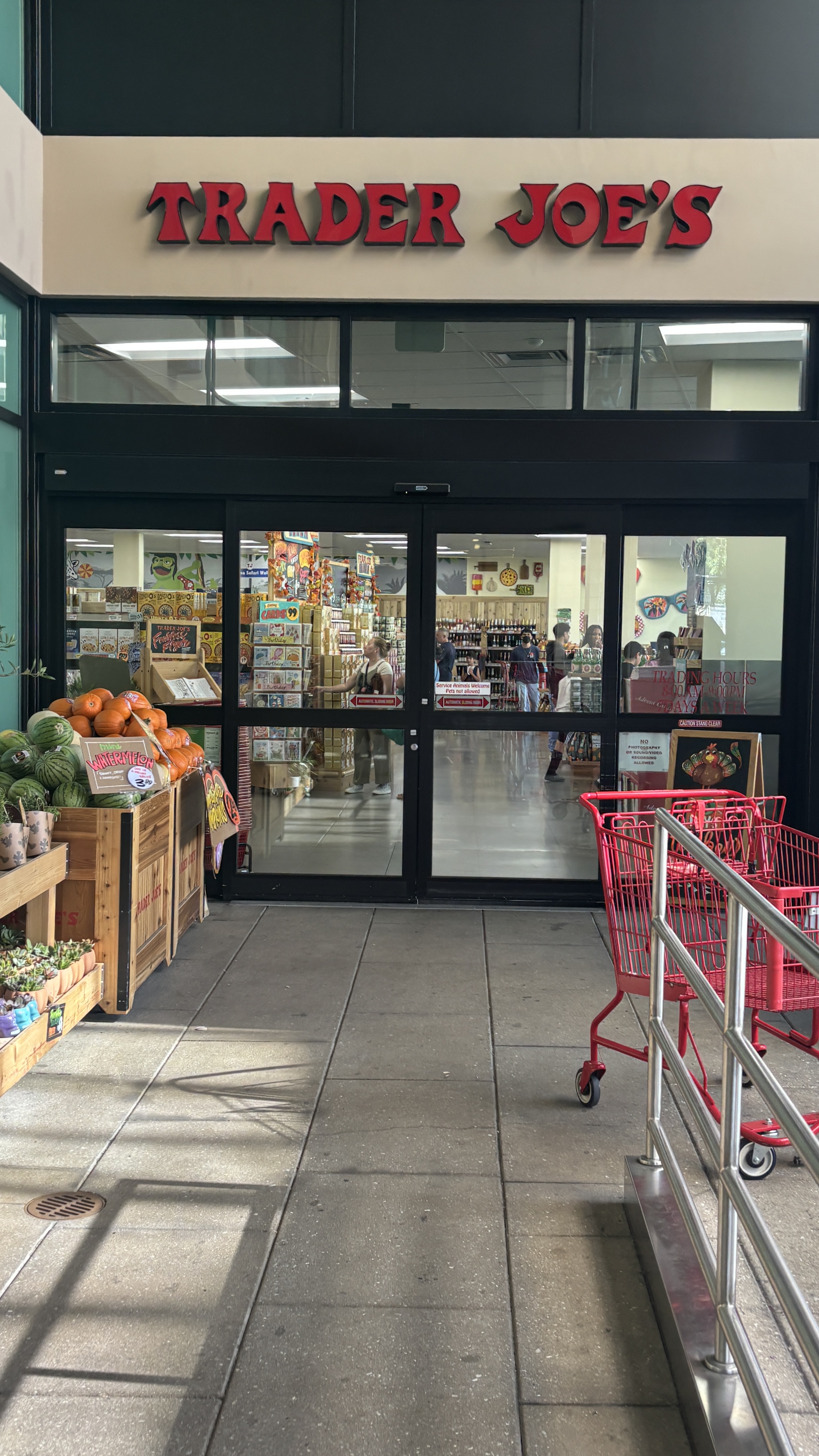 Exterior entrance of a grocery store with red signage, produce on wooden crates, and red shopping carts.