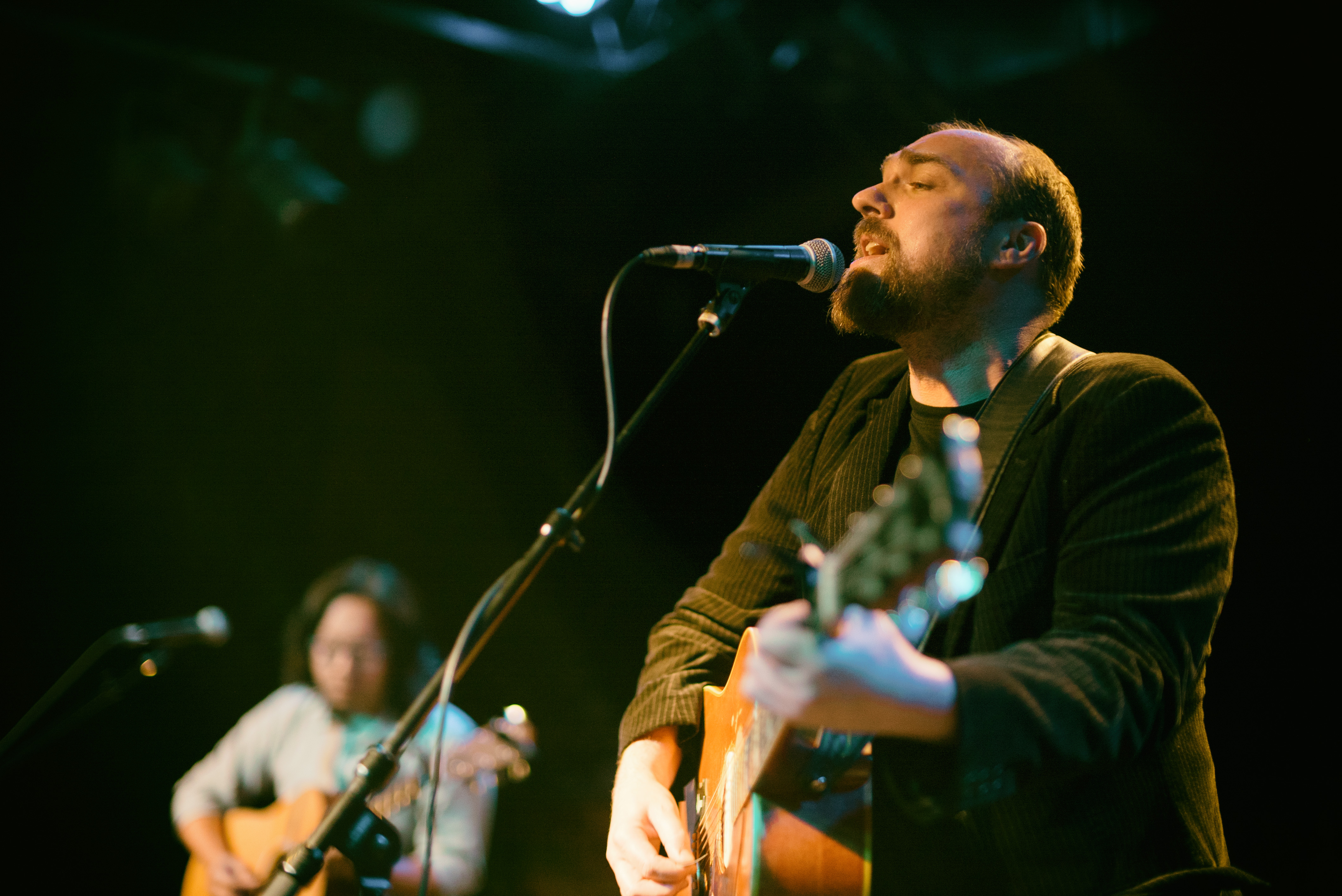 Two musicians with acoustic guitars perform on a dimly lit stage.