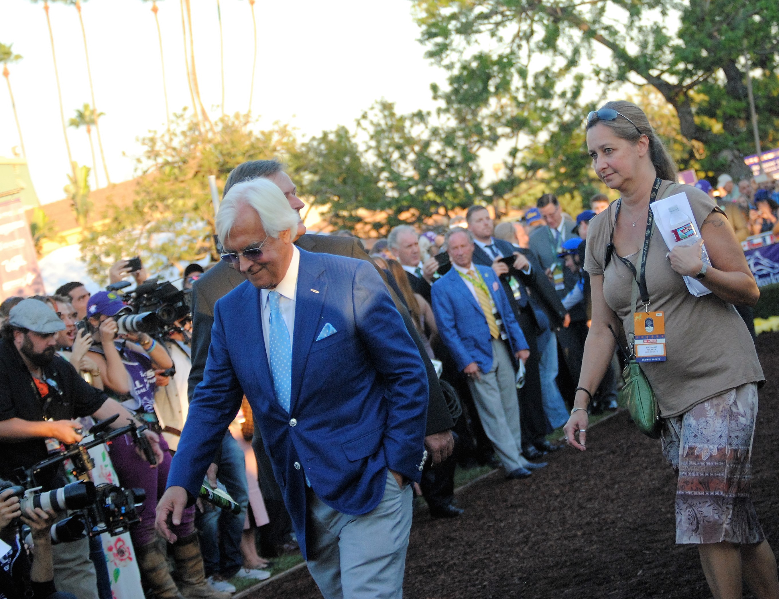 A man in a blue blazer walks through a crowd at an outdoor horse racing event.