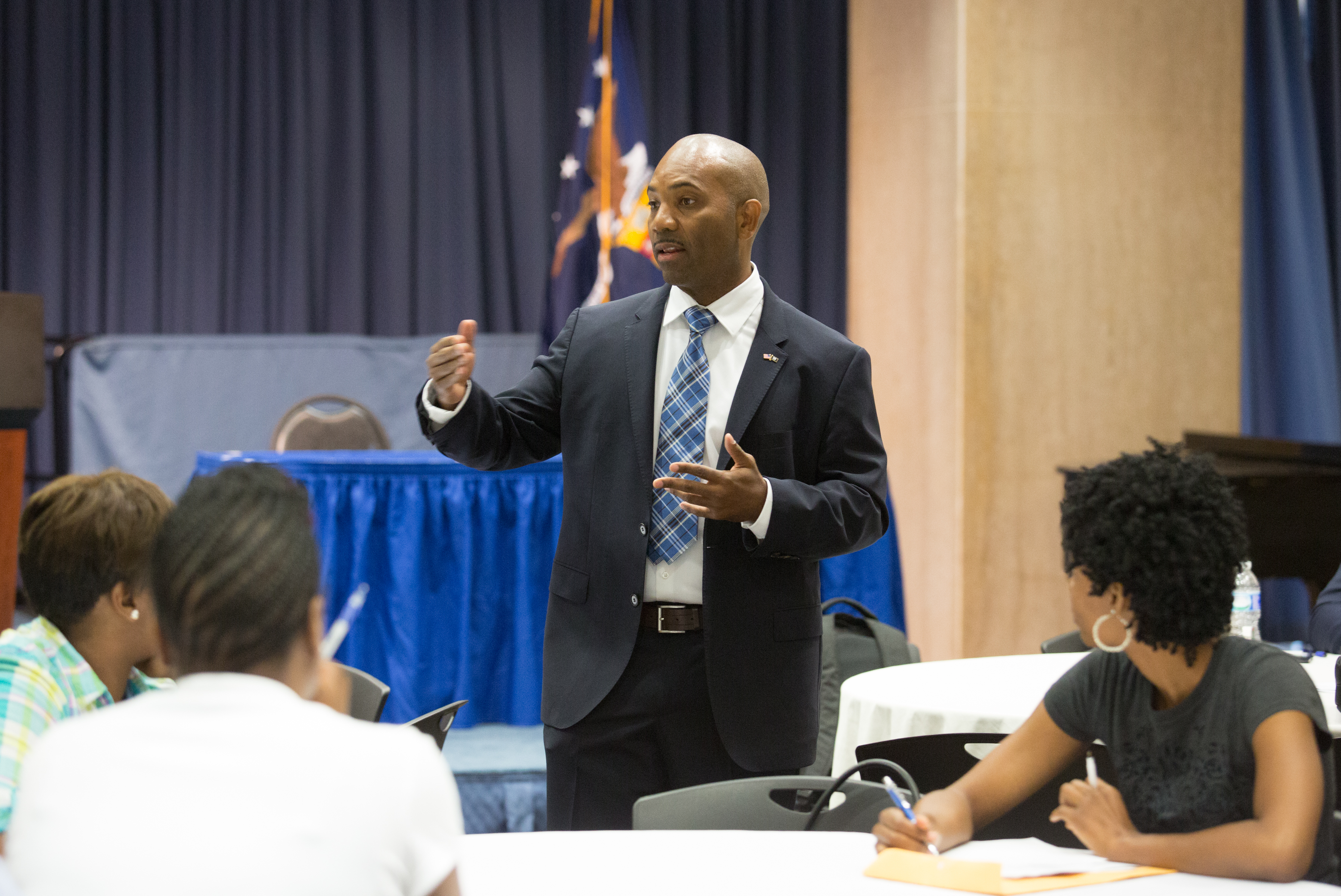 A man in a suit addresses seated candidates at a veterans employment event, American flag visible.
