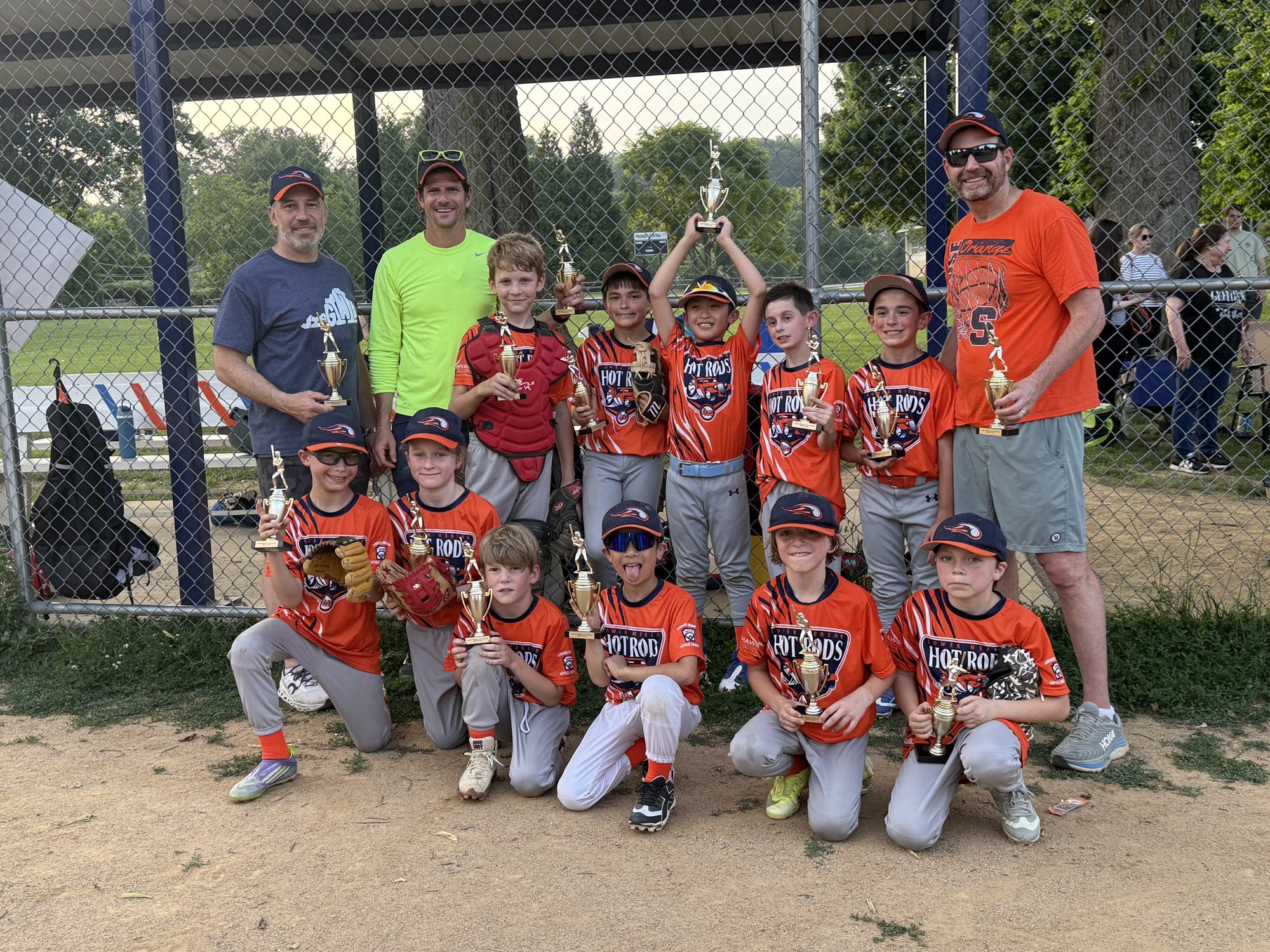 Youth baseball team in orange uniforms holds trophies in front of a chain-link fence.