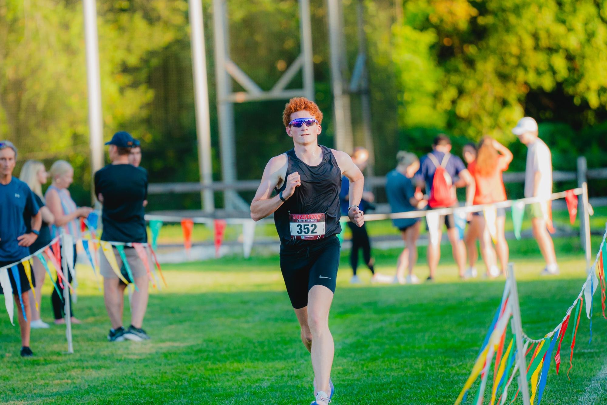 A male runner wearing a race bib sprints on a grass cross-country course with spectators behind him.