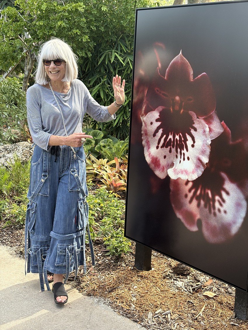 A woman with white hair stands beside a large backlit orchid photograph in a garden.