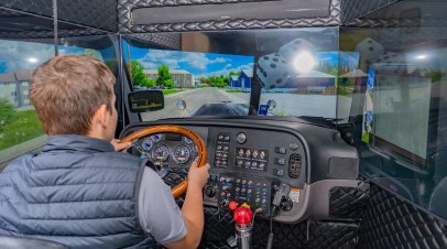 Person seated at a truck driving simulator with steering wheel and multiple screens.