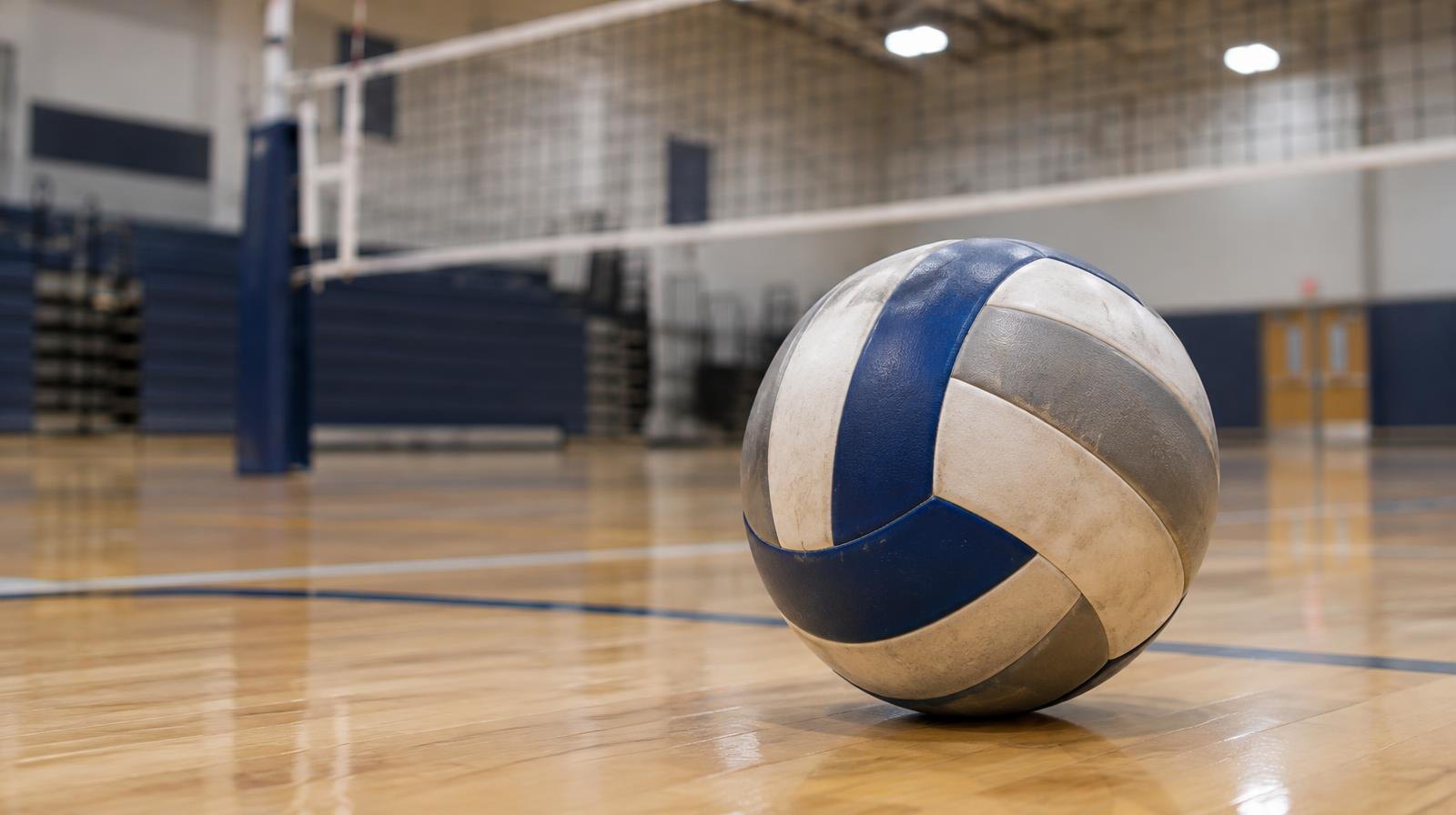 A volleyball resting on a hardwood gym floor near a volleyball net.