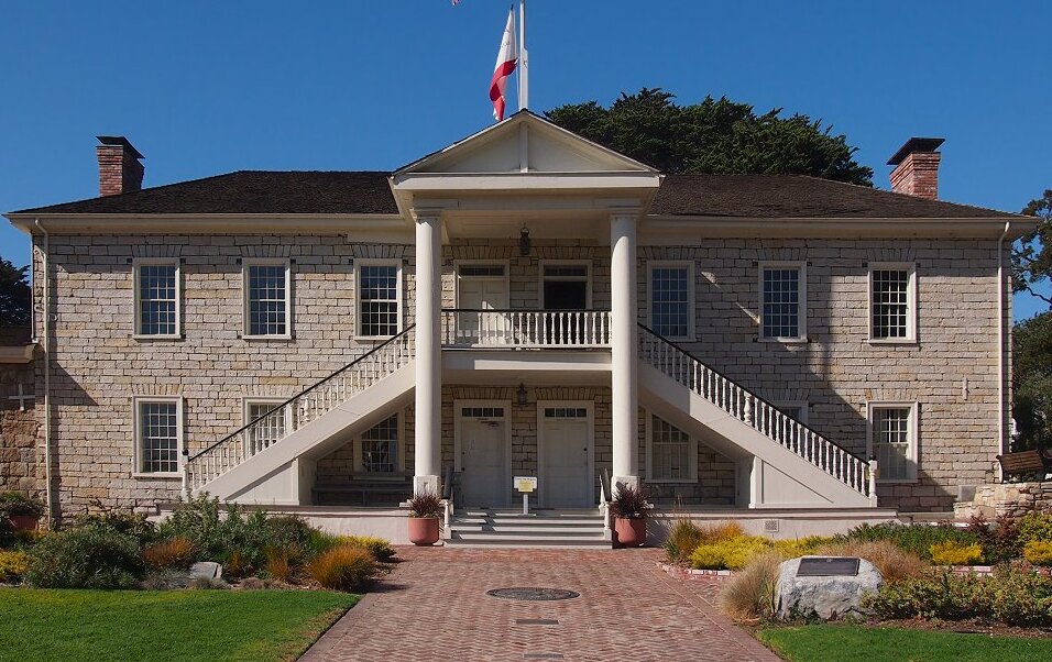 Two-story stone civic building with white columns, dual staircases, and a flagpole.