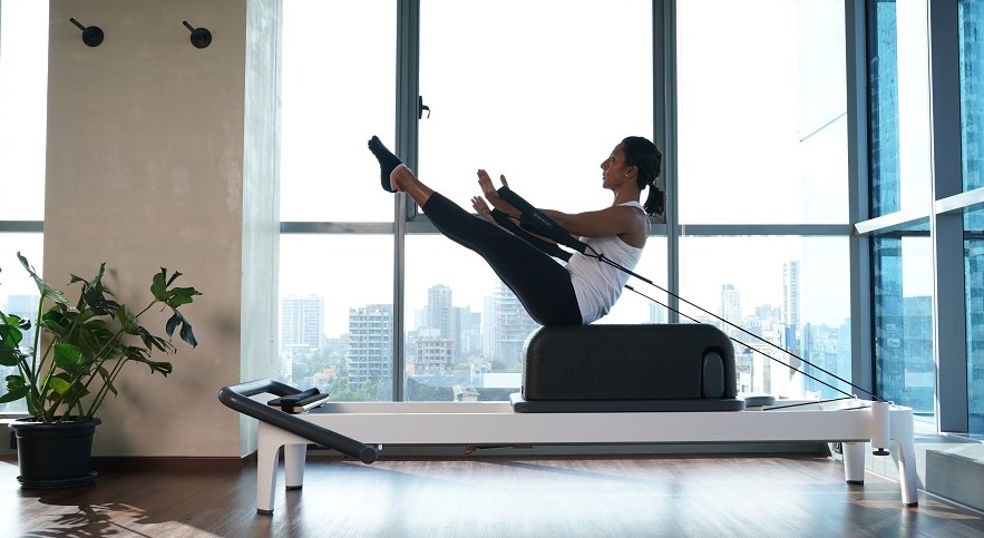 Woman exercising on a Pilates reformer machine in a bright studio with large windows.