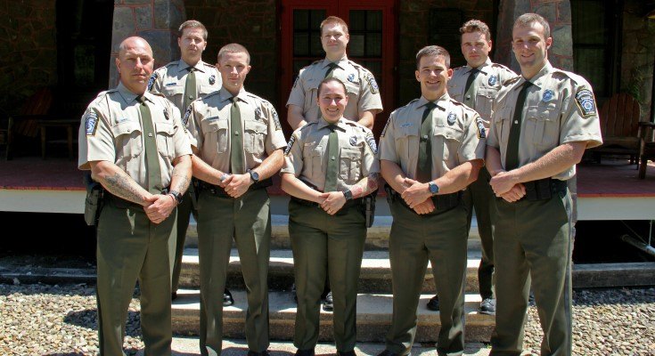 Eight uniformed conservation officers pose in two rows outdoors in front of a building.