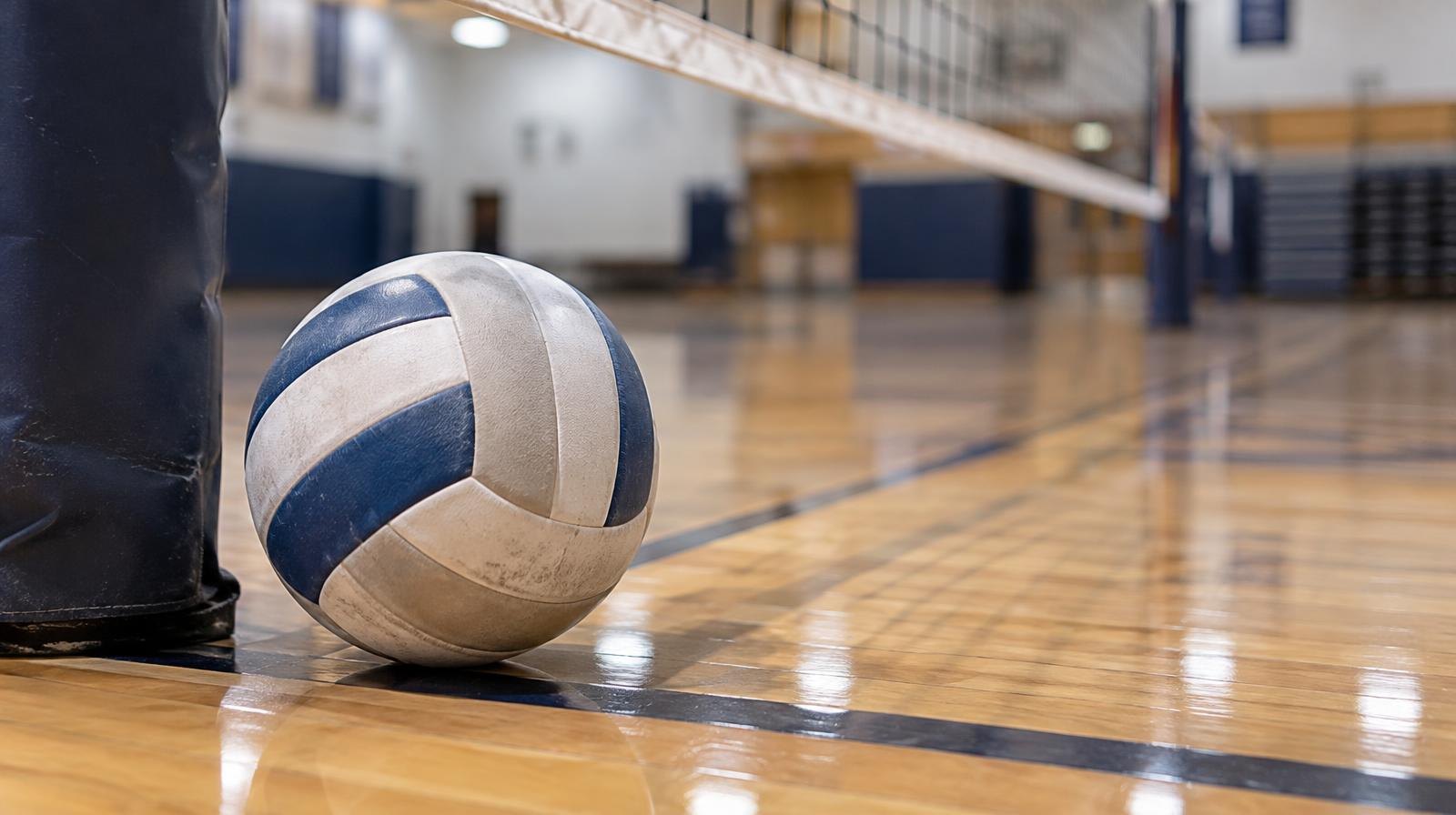 A volleyball resting on a hardwood gym floor next to a net post.