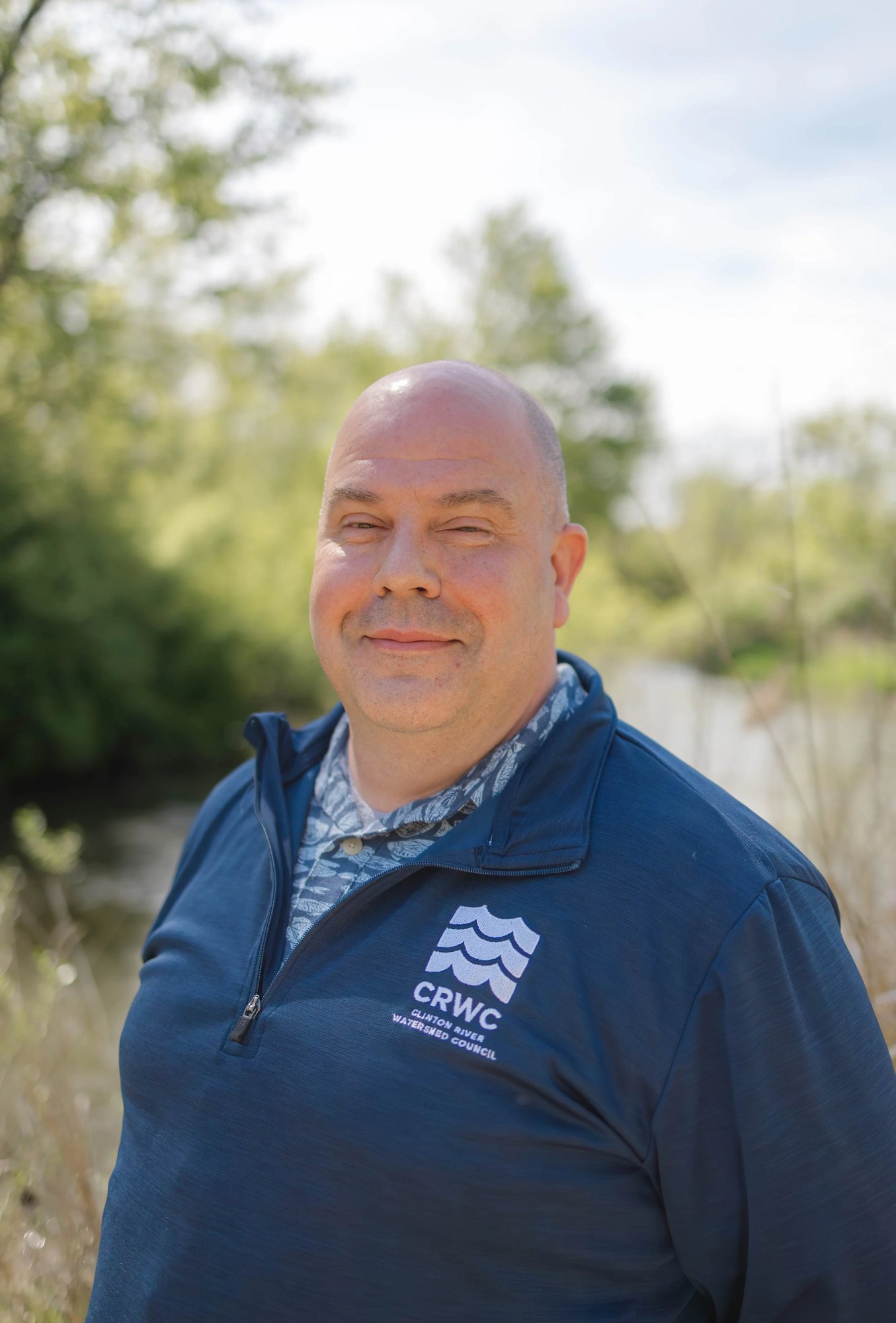Person wearing a CRWC jacket standing outdoors near a waterway with tall grasses.