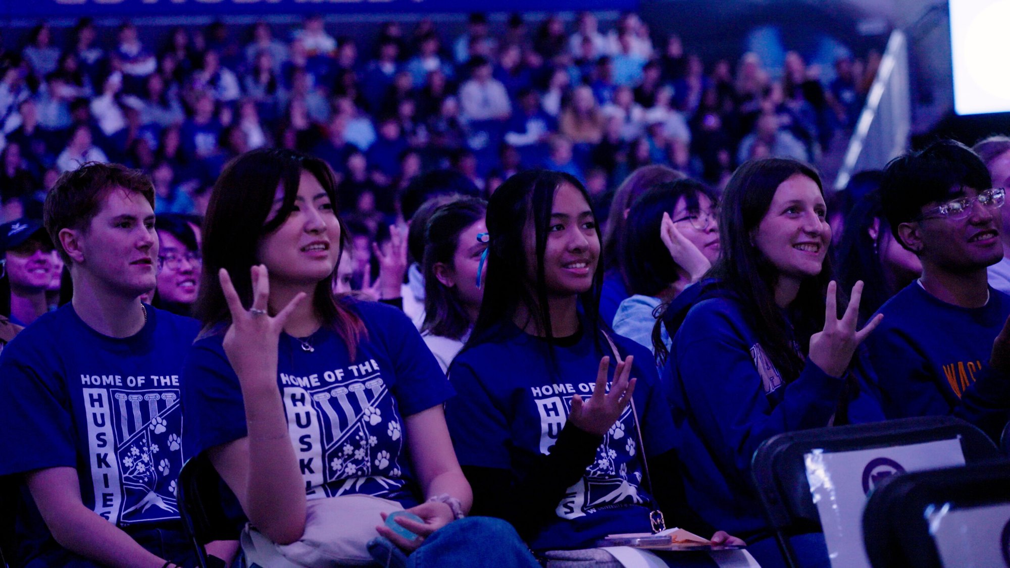 Students in a large arena audience smile and make hand gestures at a university convocation.