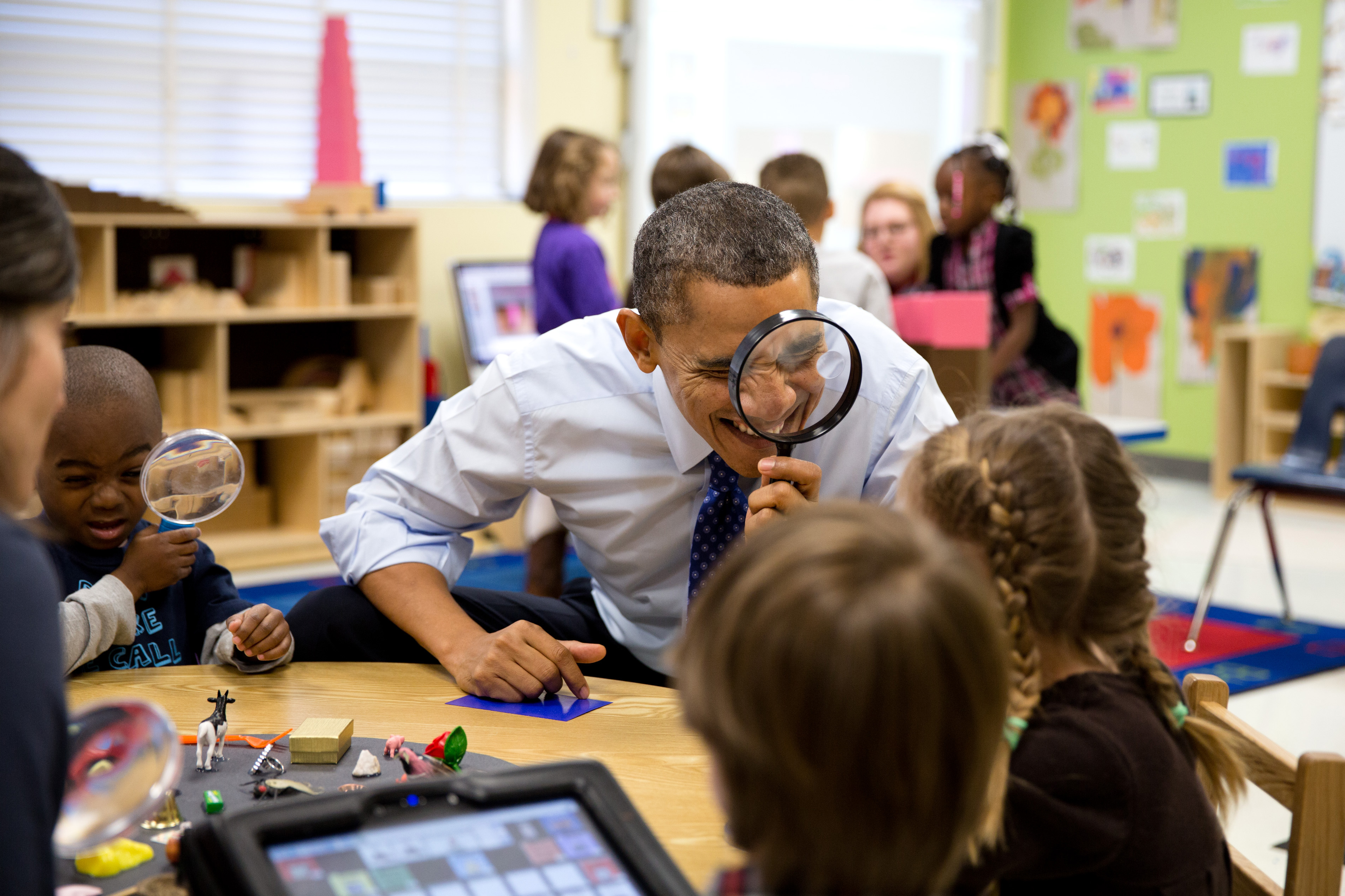 An adult holds a magnifying glass to his face while leaning over a table with young children in a colorful preschool classroom.