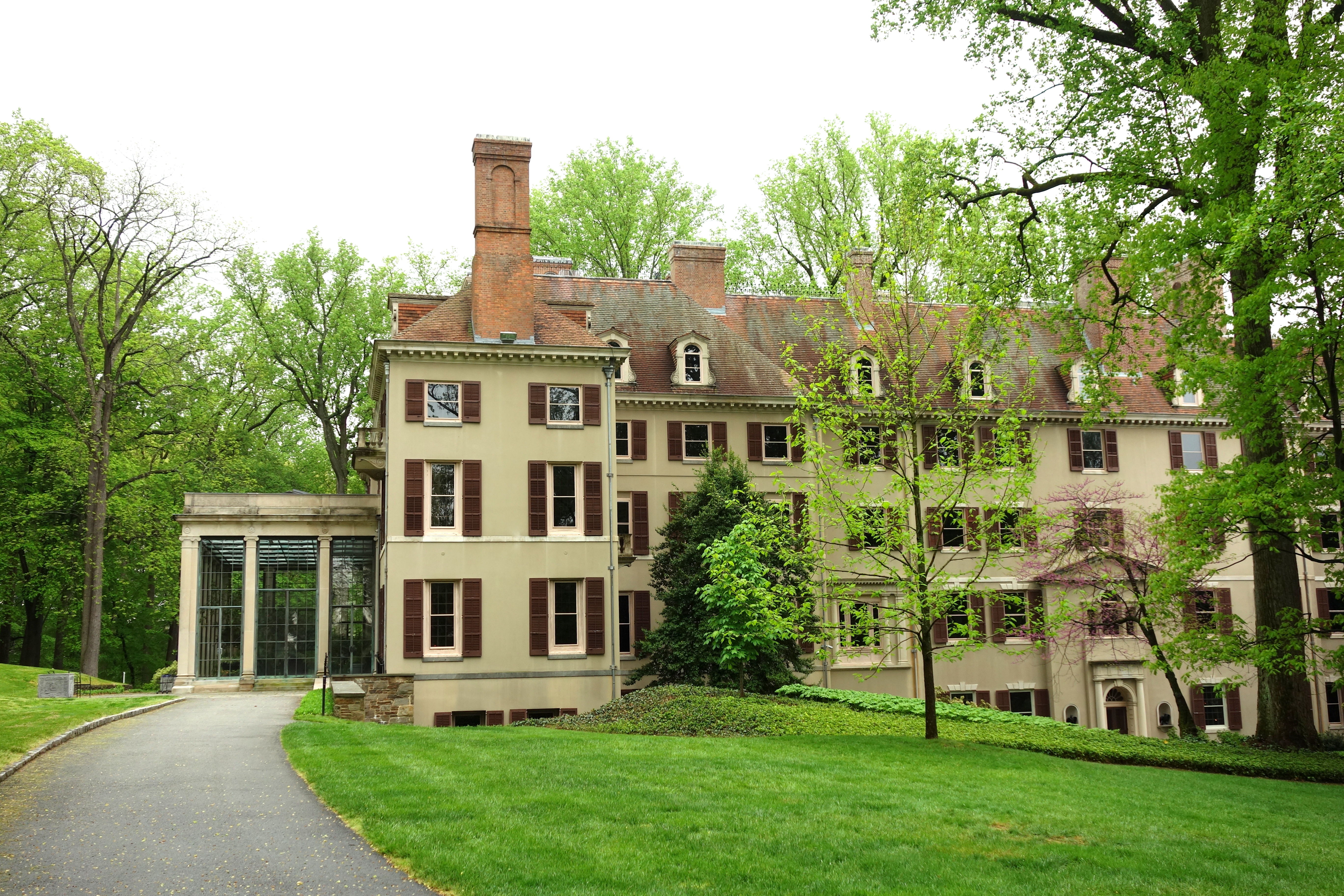 Large historic mansion with dark shutters, a glass addition, and mature trees on a green lawn.