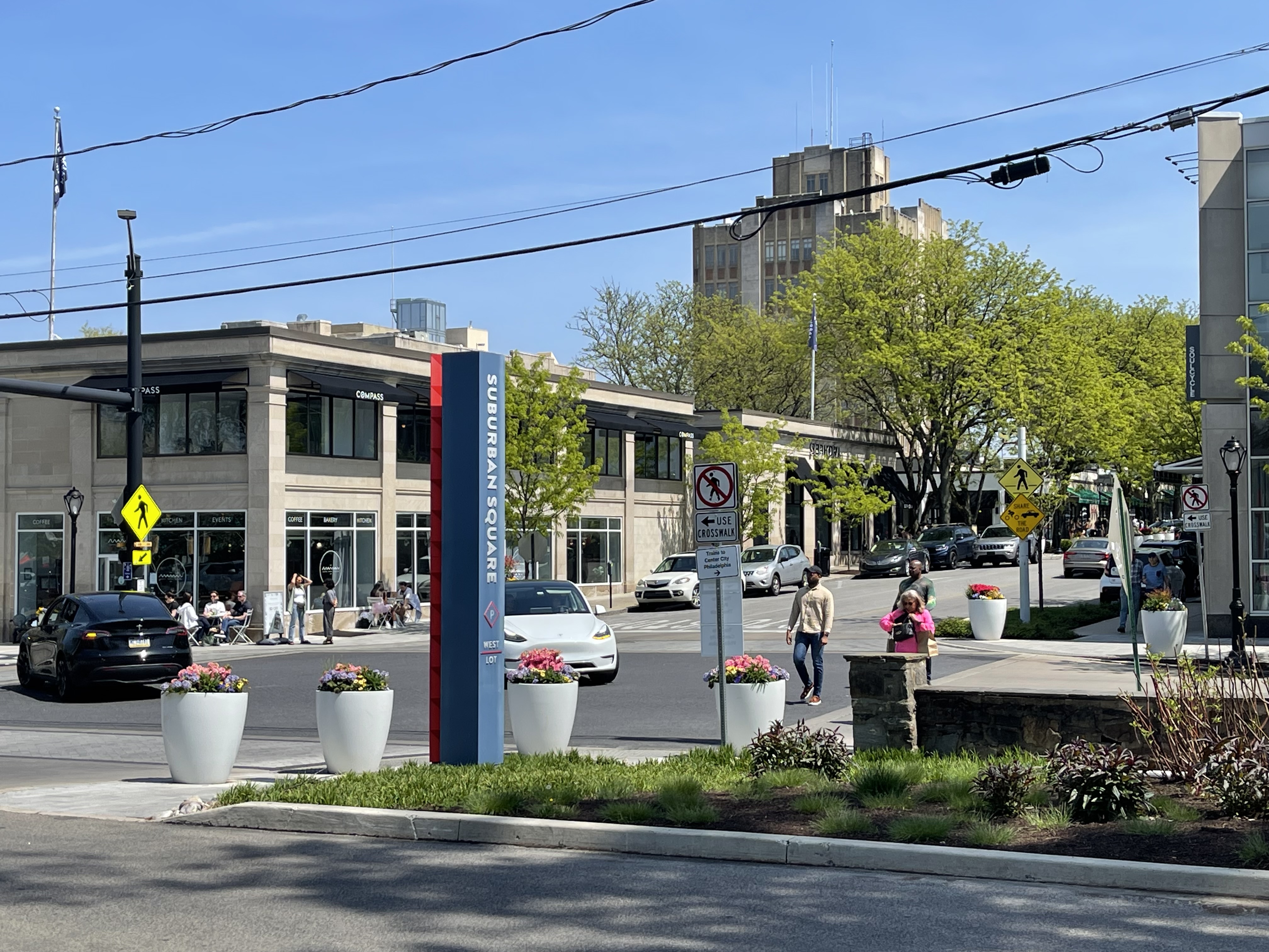 Pedestrians walk past a blue directory sign and flower planters at an outdoor shopping center.