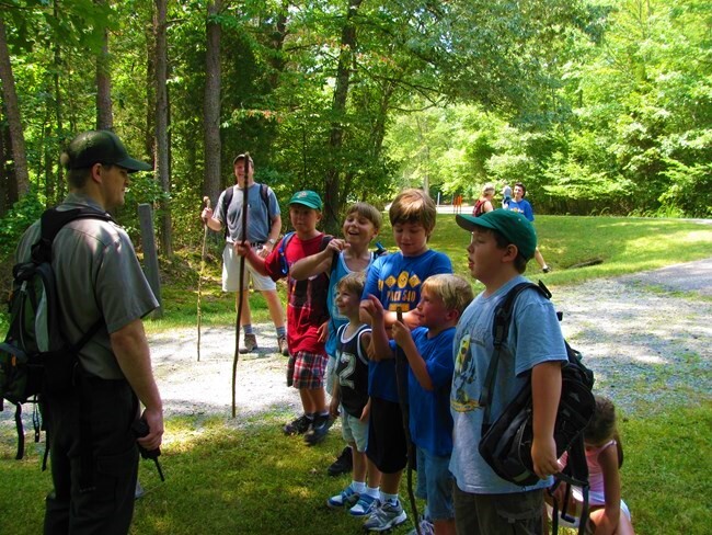 An adult guide leads a group of children along a wooded gravel trail.