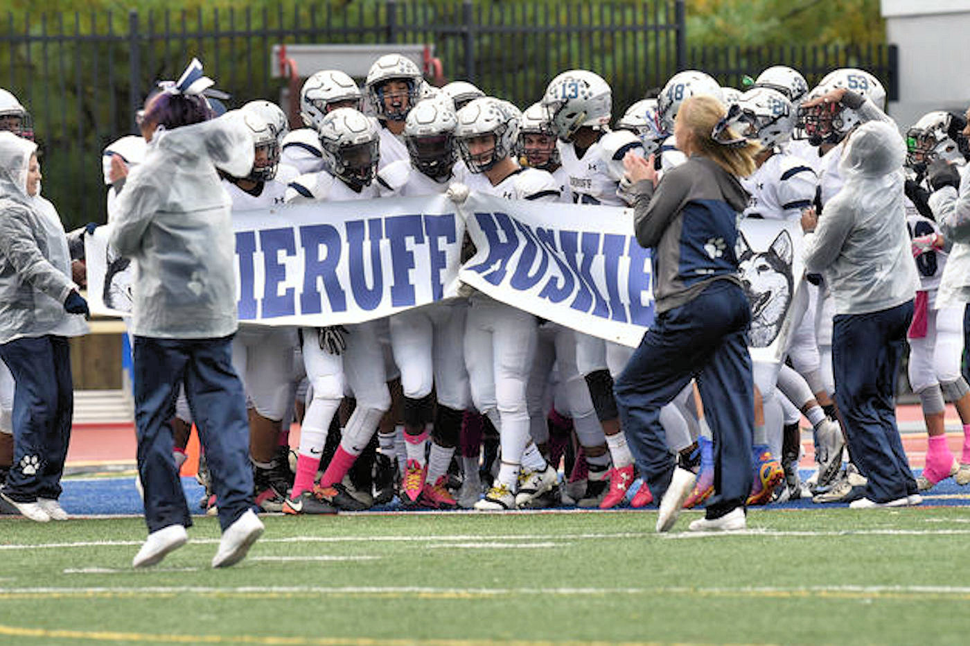 High school football players in white uniforms burst through a paper banner onto a turf field.