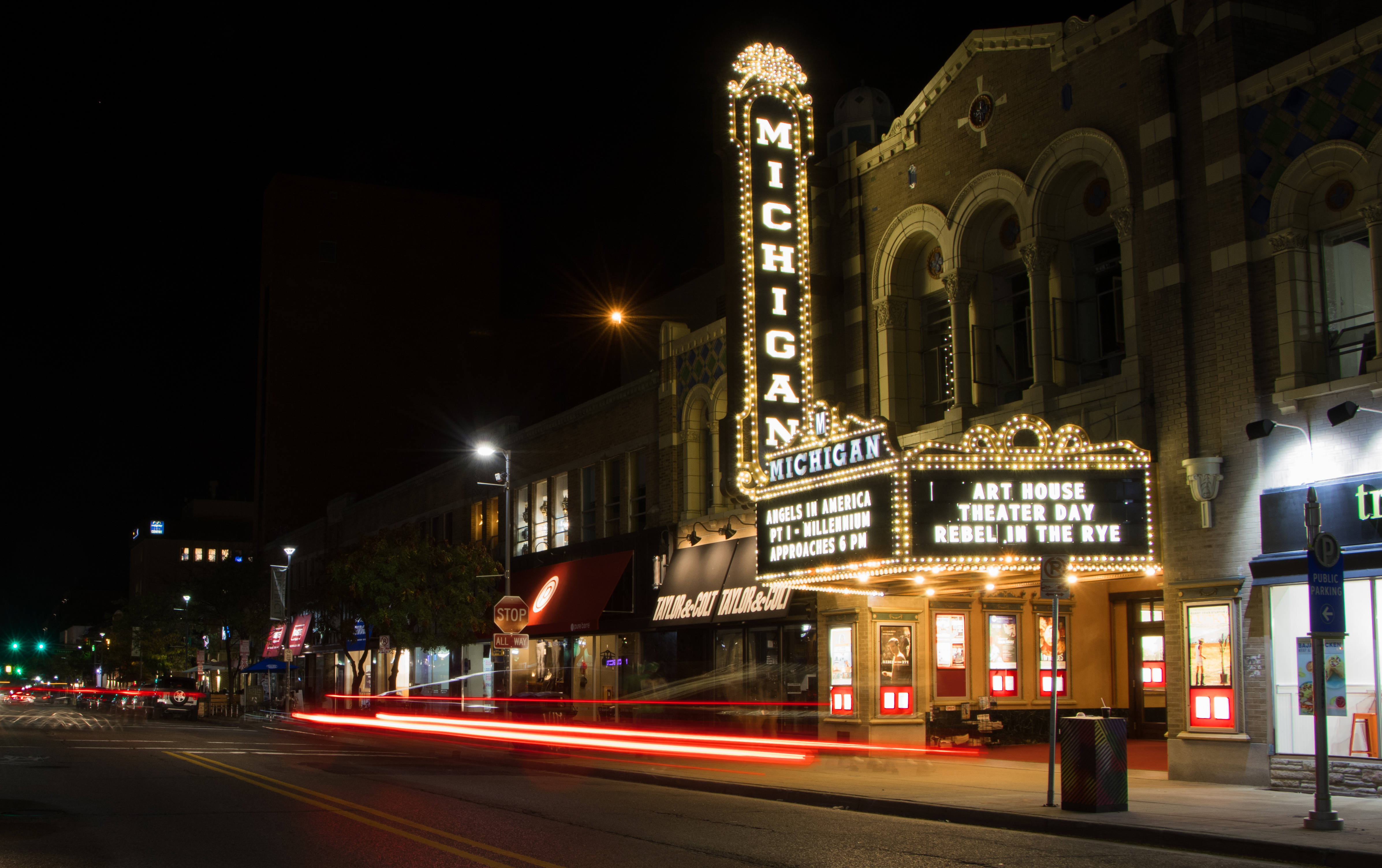 Illuminated theater marquee at night with light trails from passing vehicles on the street below.