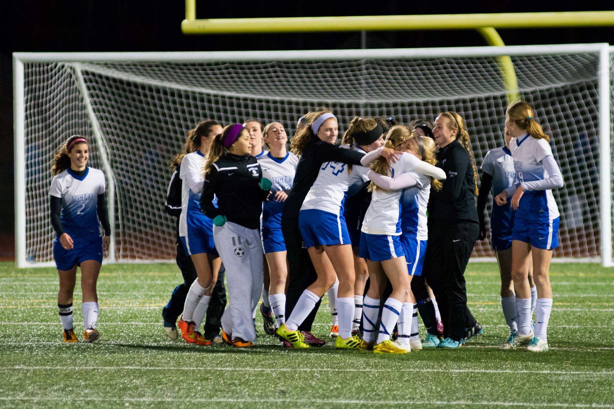 Girls soccer players in blue and white uniforms celebrate near a goal at night.