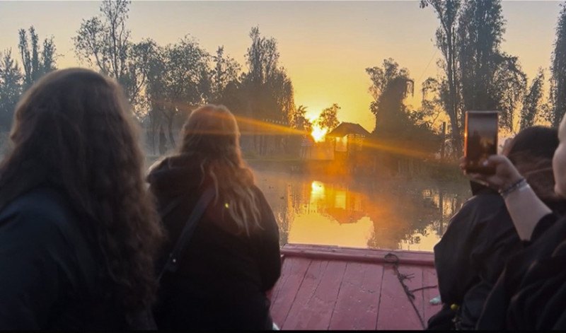 Several people seated on a red flat-bottomed boat watch a golden sunrise over a misty lake.