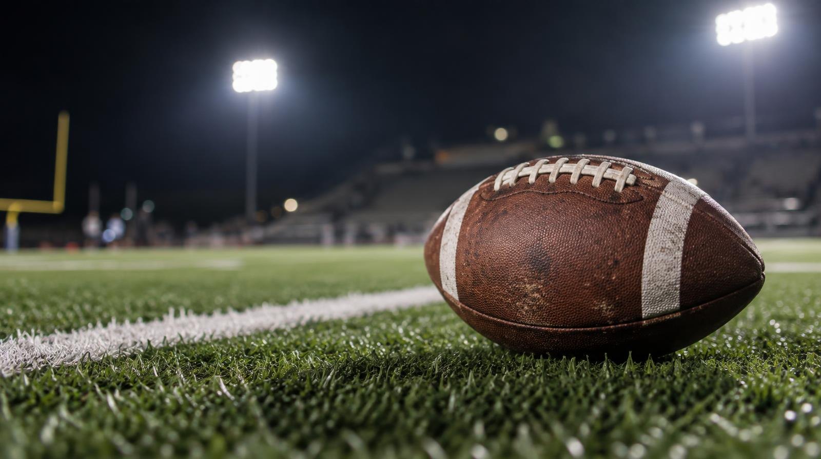 An American football resting on green artificial turf beside a white yard line under stadium lights.