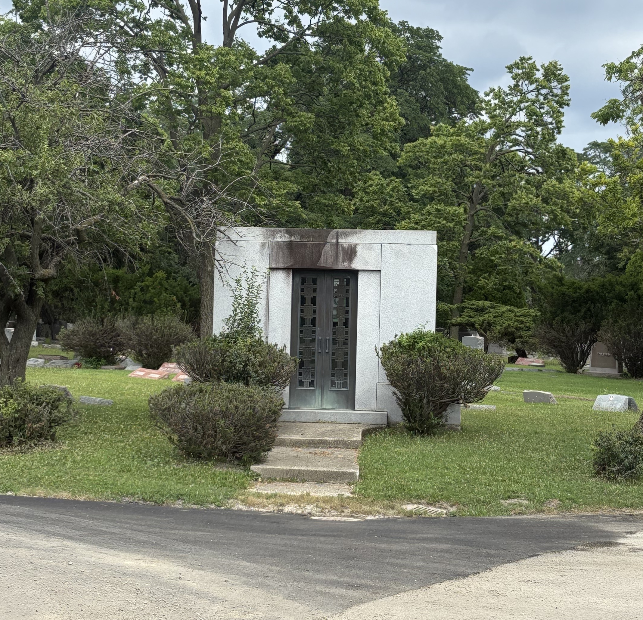 Stone mausoleum with metal doors flanked by trimmed shrubs in a cemetery.