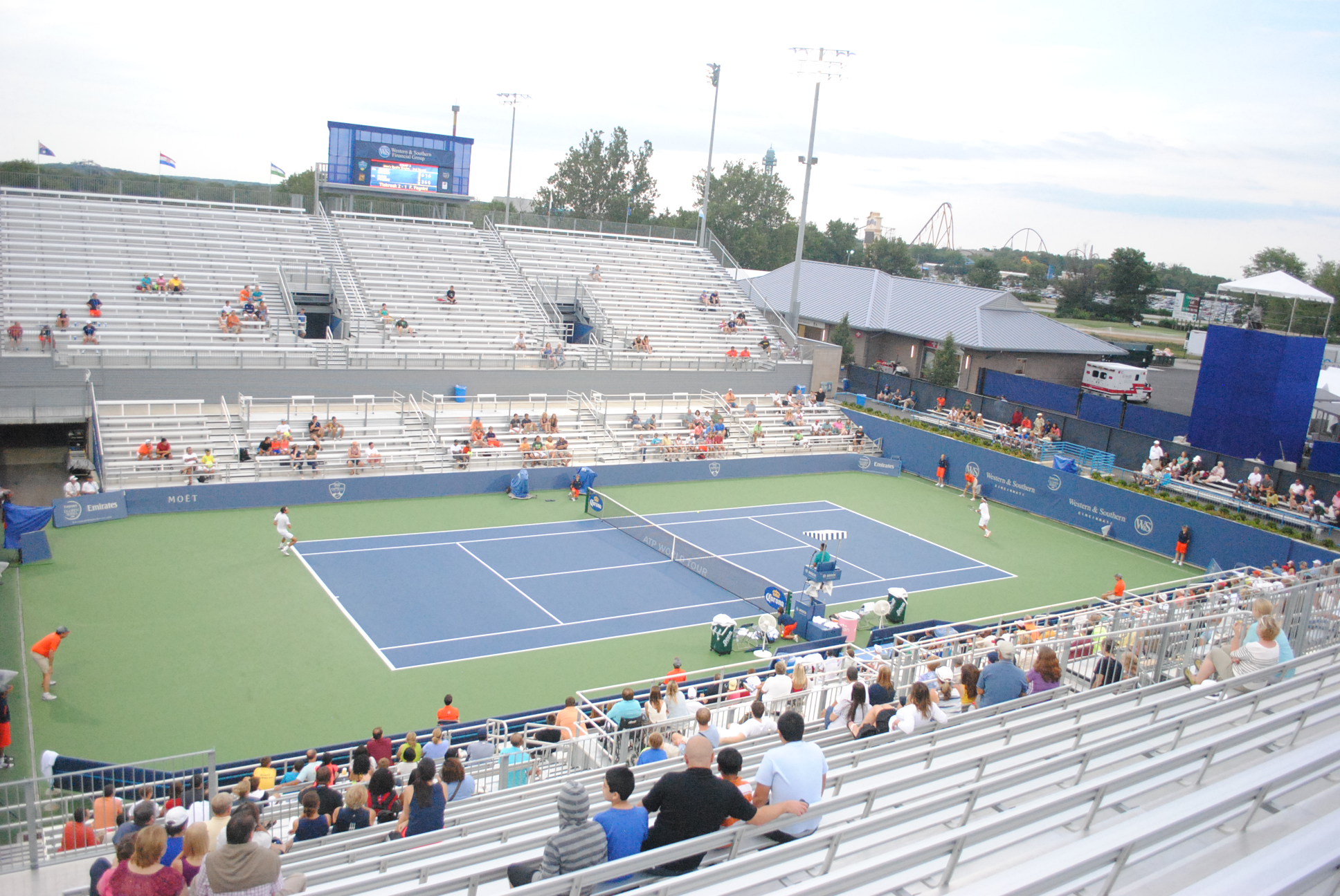 Outdoor tennis match on a blue hard court with spectators in the stands and a scoreboard in the background.