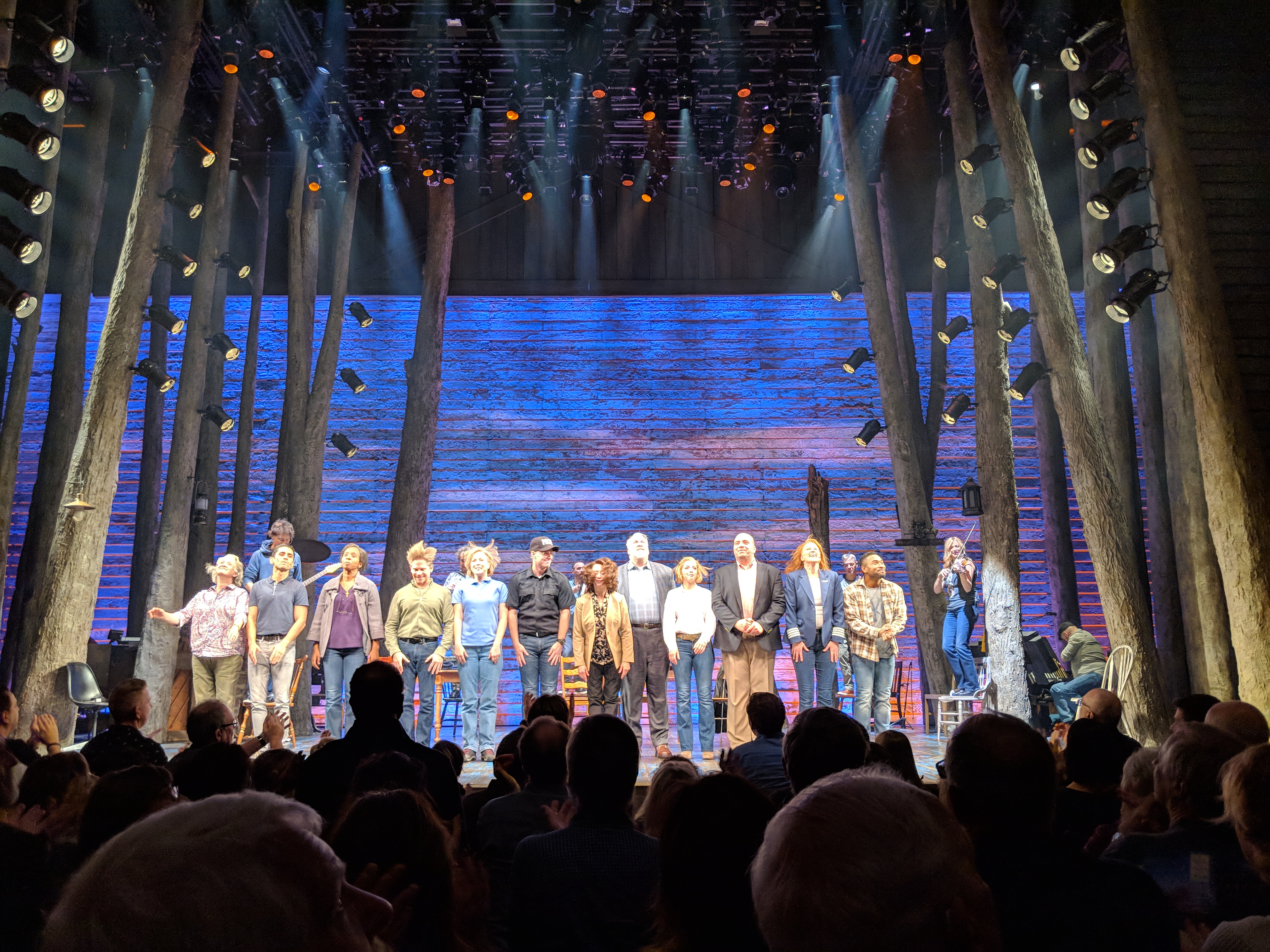 Large ensemble cast bowing on a wooden-plank stage surrounded by tall tree-trunk columns.