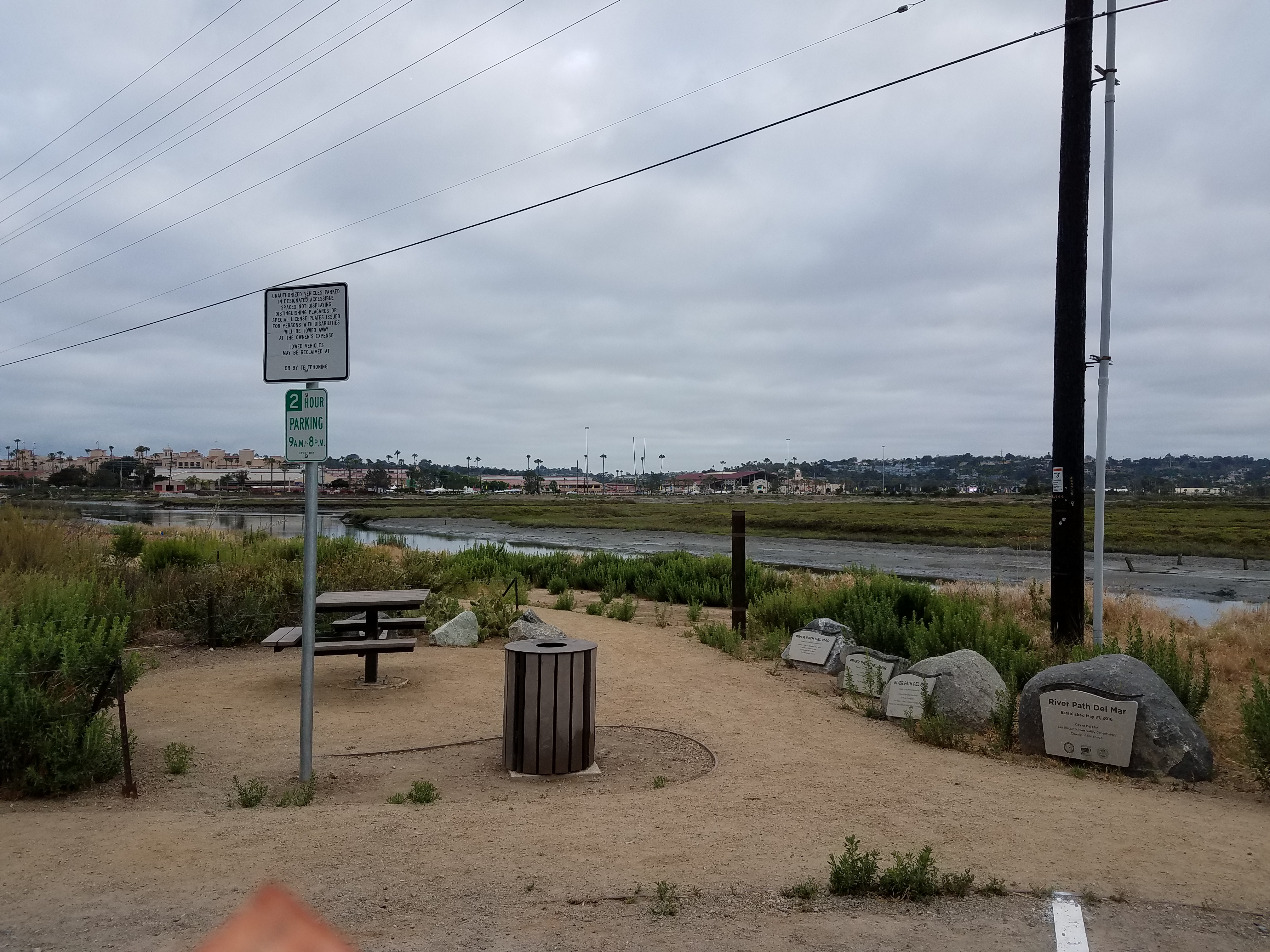 Sandy trailhead with informational plaques and a lagoon waterway in the background.