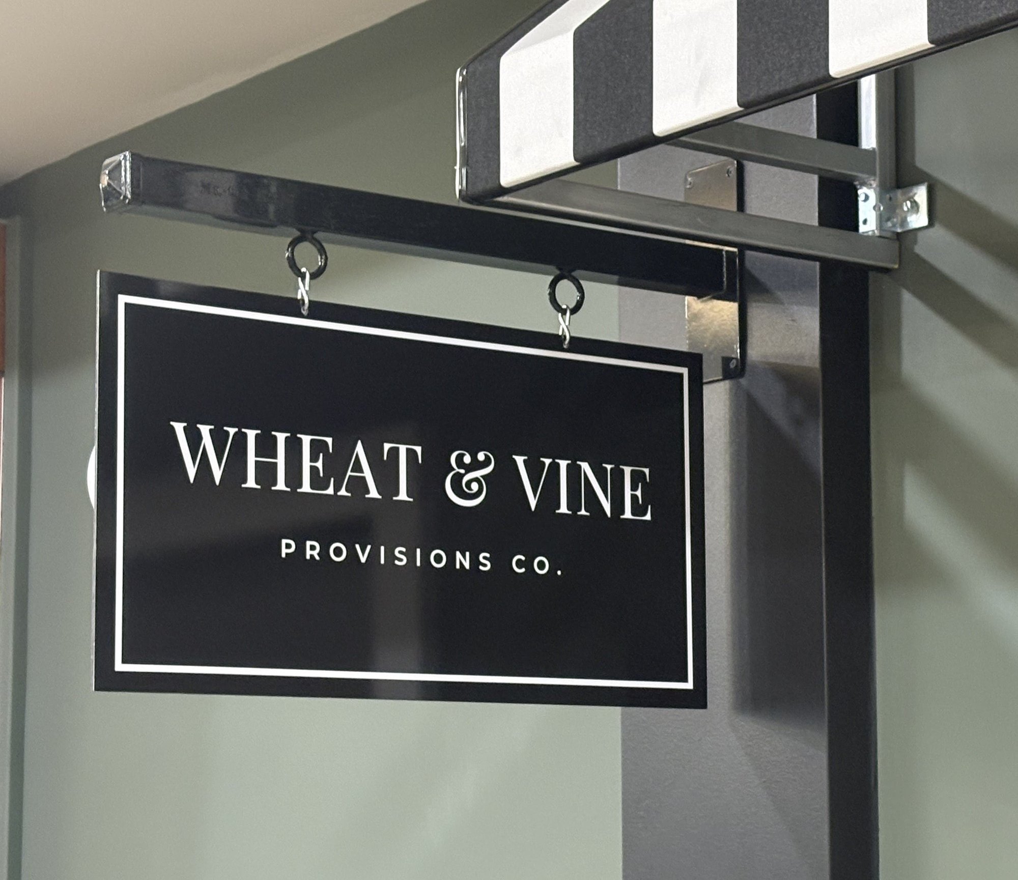 Black hanging sign reading 'Wheat & Vine Provisions Co.' outside a storefront with a striped awning.