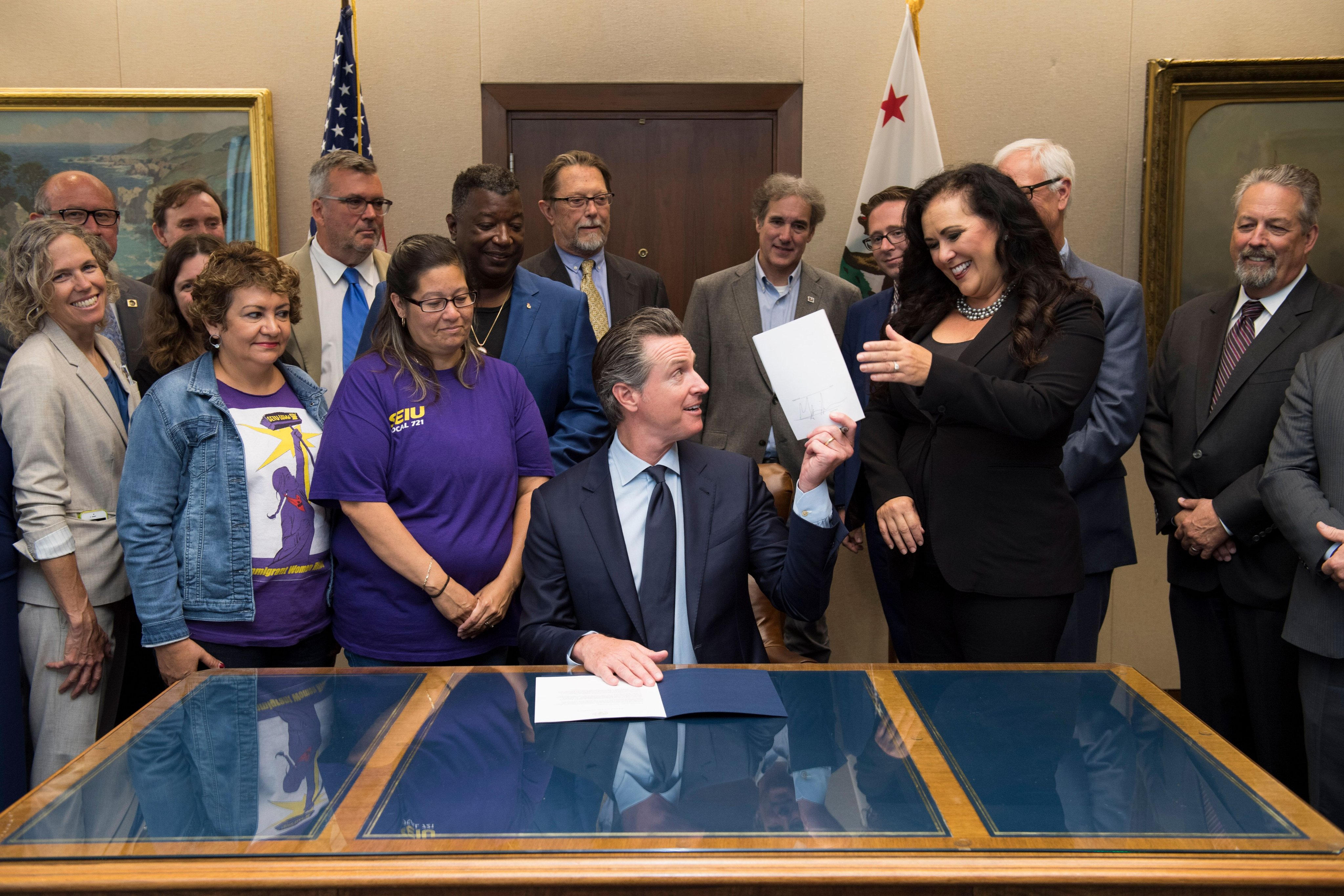 Governor seated at a signing desk surrounded by officials with US and California flags behind them.
