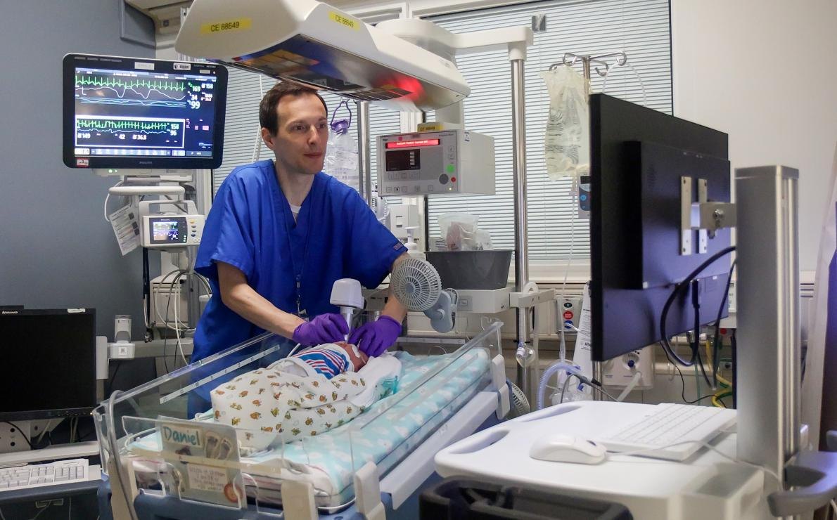 Healthcare worker in blue scrubs examining a premature infant inside a NICU incubator surrounded by monitors.